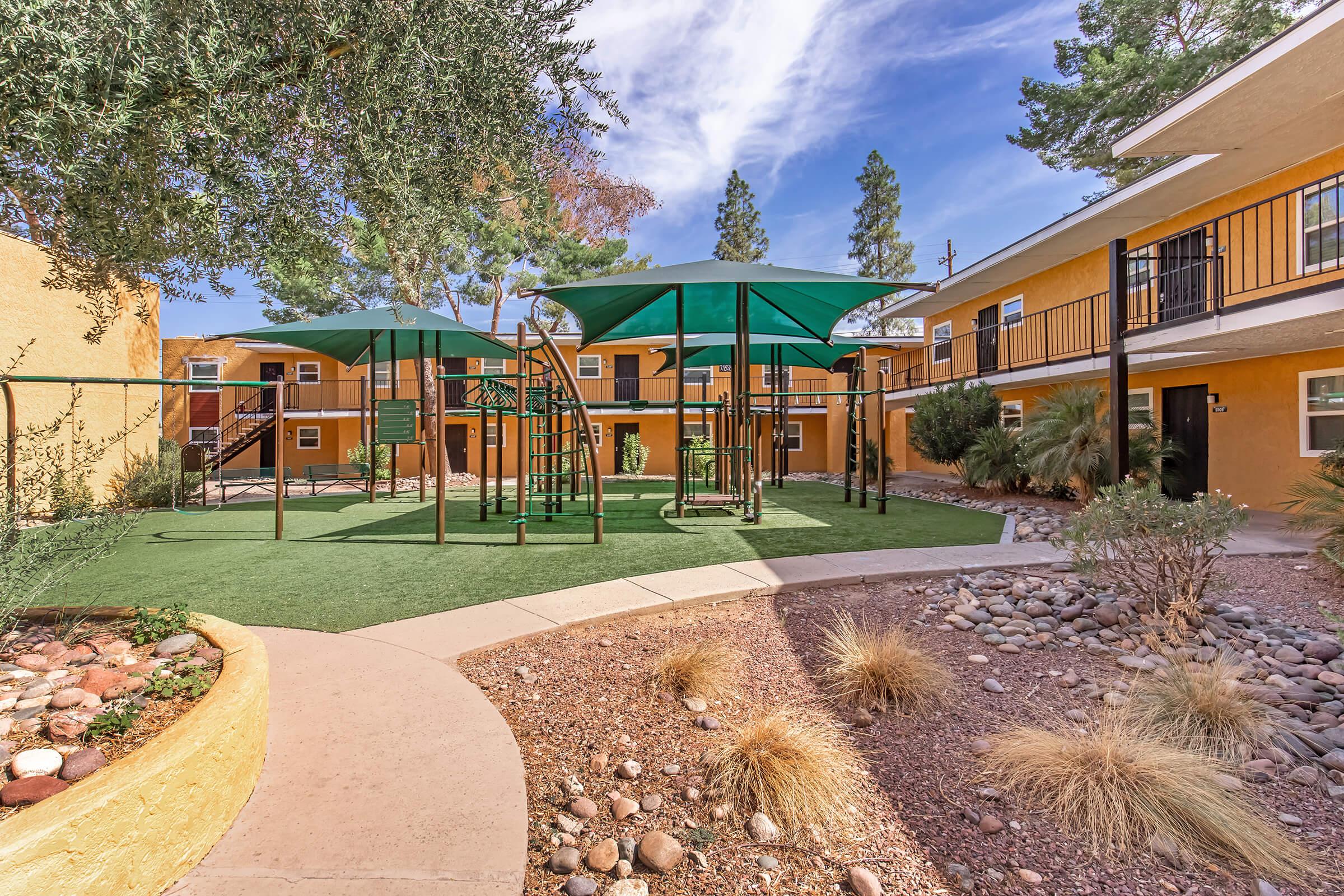 A playground area with green equipment and shaded structures, set in a landscaped courtyard surrounded by buildings. The space features artificial turf, gravel landscaping, and plants, with clear blue skies above.