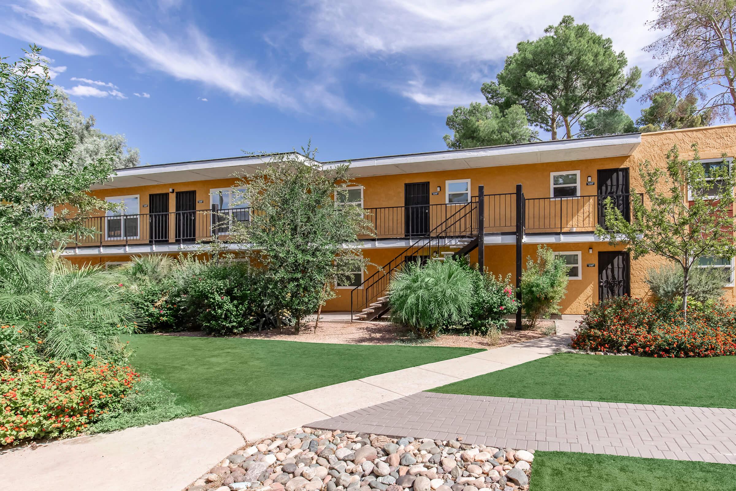 A two-story apartment building with a yellow exterior, surrounded by green lawns and colorful flowering plants. The building features black railings on the porch and trees in the background under a clear blue sky with some clouds. A pathway made of stone leads through the landscaped area.