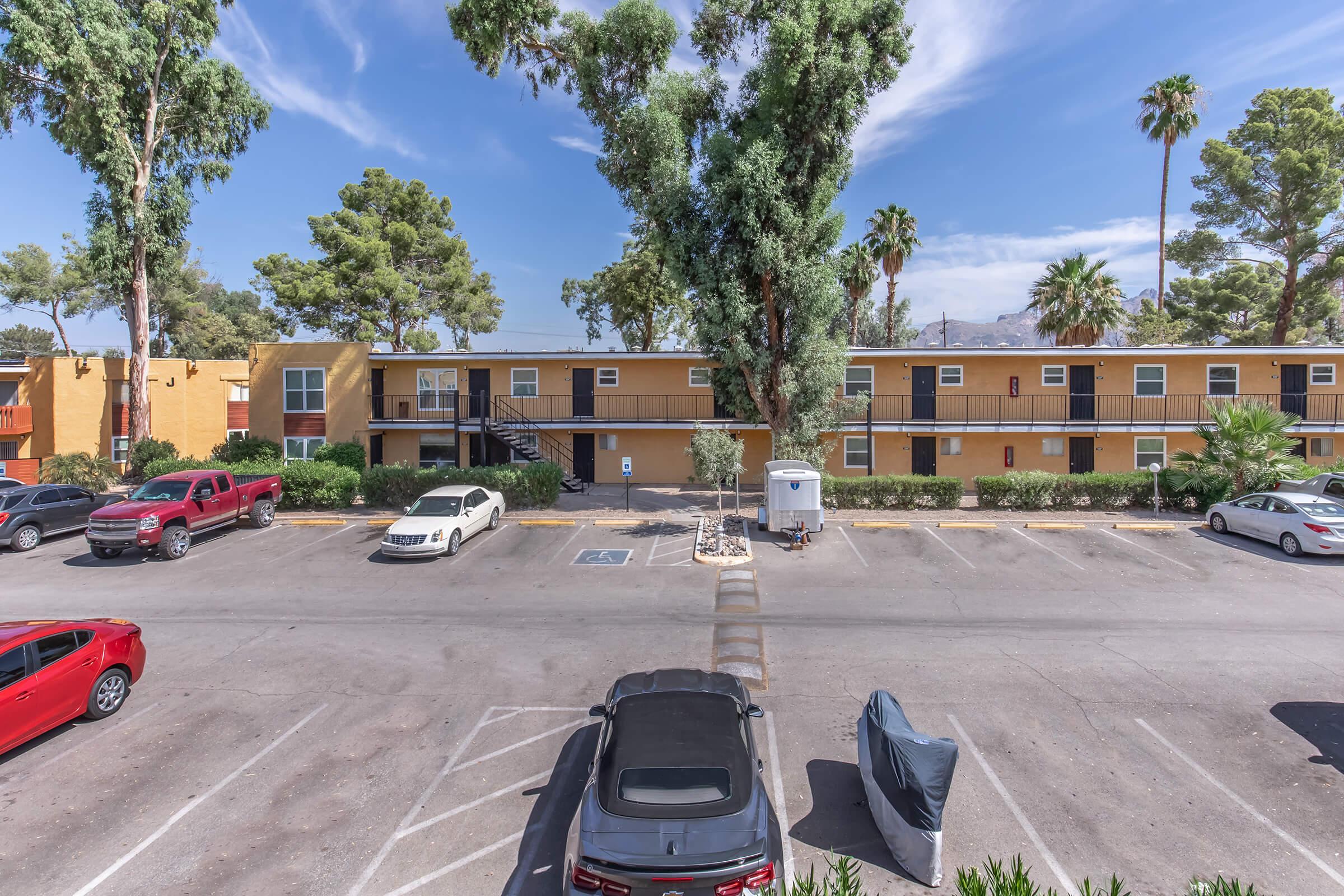View of a sunny parking lot with several parked cars in front of a two-story apartment building. The building features balconies and is surrounded by palm trees and other vegetation. In the background, there are mountains visible against a clear blue sky.