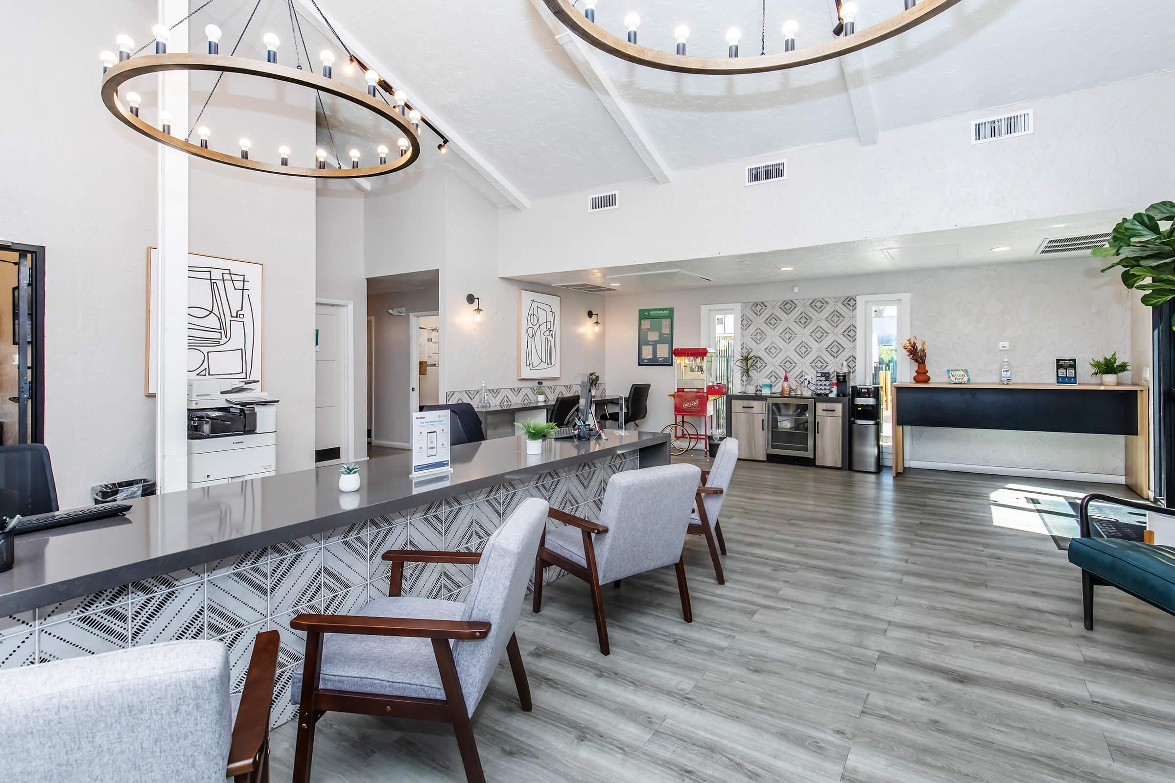 A modern, well-lit reception area featuring a sleek countertop, stylish chairs, and contemporary decor. Large windows allow natural light to fill the space, complemented by a statement chandelier. The background includes a kitchenette with modern appliances and decorative elements like plants and artwork.