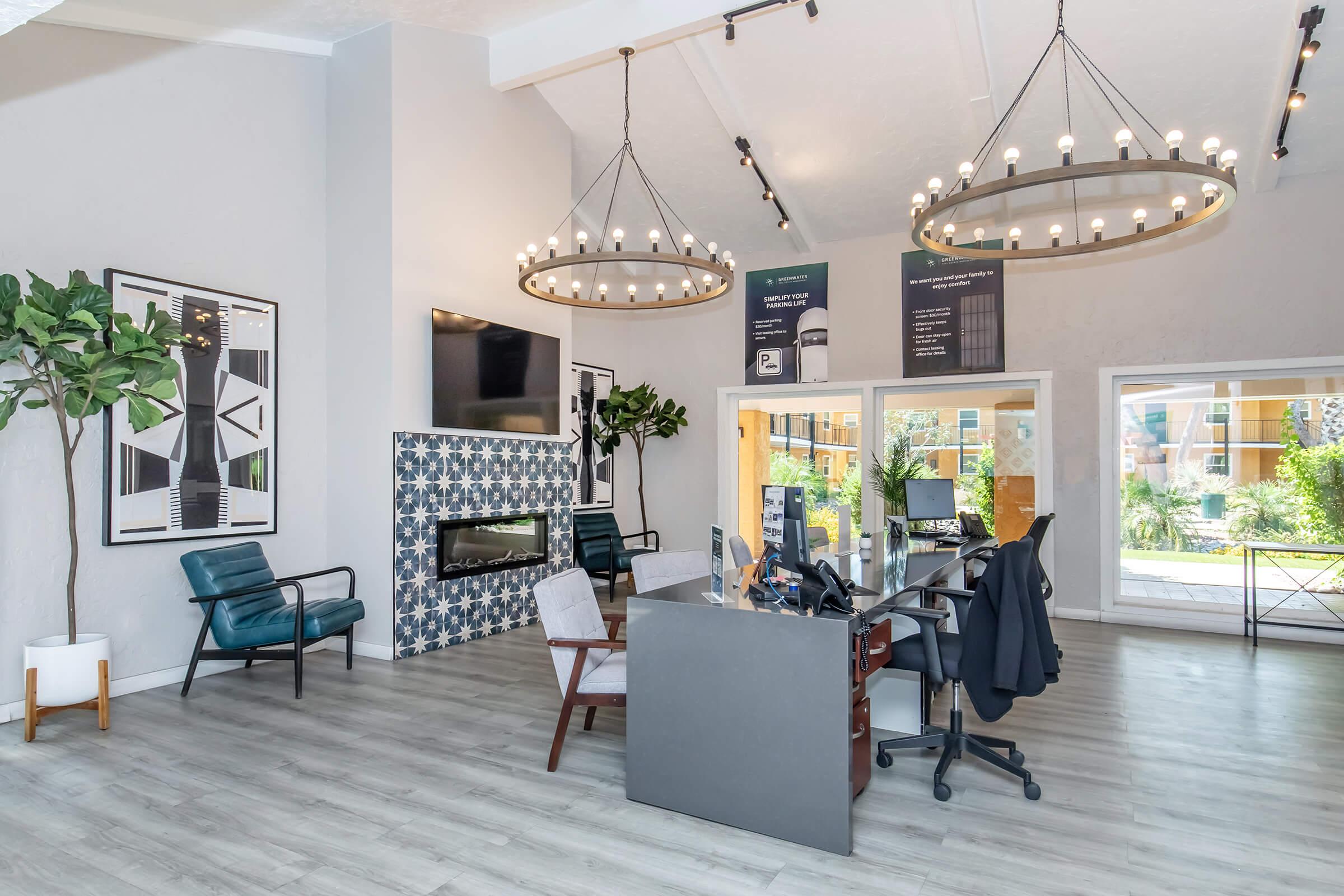 A modern lobby area featuring high ceilings with large chandeliers, a stylish seating arrangement with colorful armchairs, a decorative fireplace, and large windows offering views of greenery outside. The space includes a reception desk and contemporary artwork on the walls.