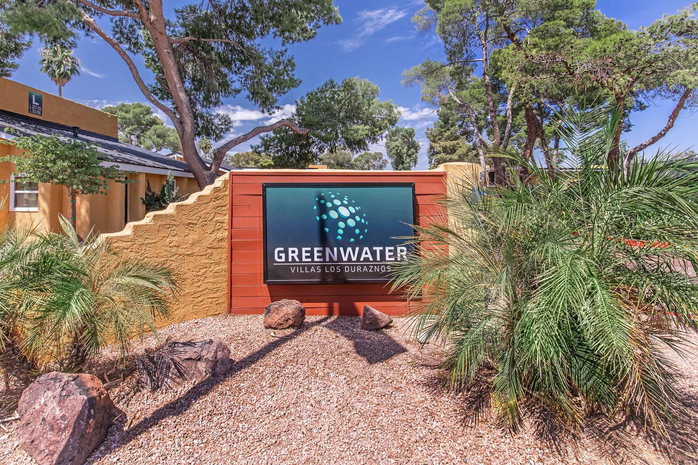 A colorful sign reading "GREENWATER VILLAS LOS DURAZNOS" surrounded by palm trees and desert landscaping. The sign is mounted on a textured wall, with rocks and gravel in the foreground. The scene is bright and sunny, showcasing a welcoming entrance to a villa property.