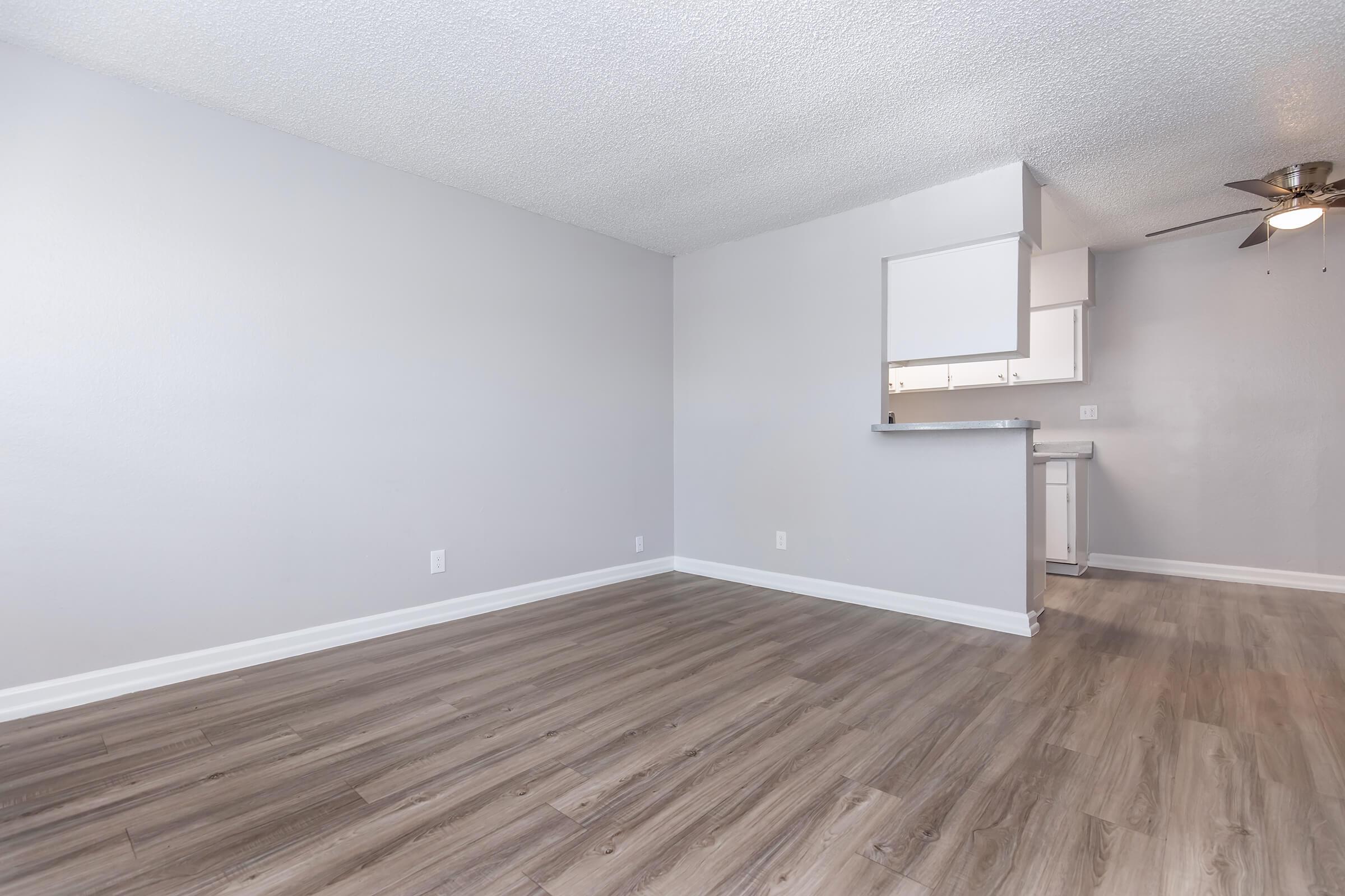 Empty room with light gray walls and new wood-like flooring. A ceiling fan is visible in the upper right corner. In the background, a small kitchen area is partially visible, featuring a countertop and cabinets. The space appears clean and well-lit, suggesting a modern and minimalist design.
