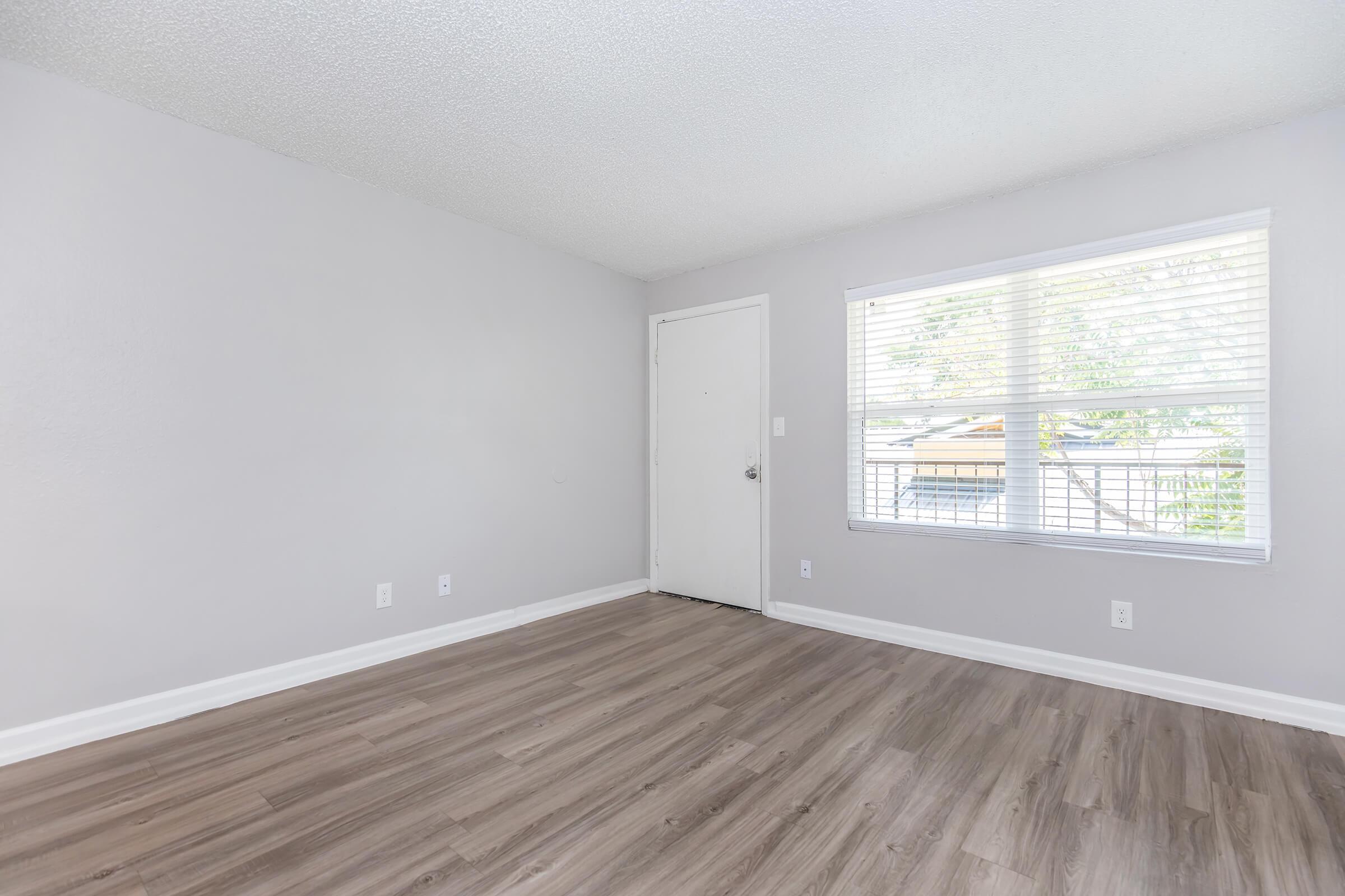 Empty room with light gray walls and wooden flooring, featuring a window with blinds that lets in natural light. A plain door is visible on one wall, and there are no furnishings or decorations. The space appears clean and bright, ideal for various uses.
