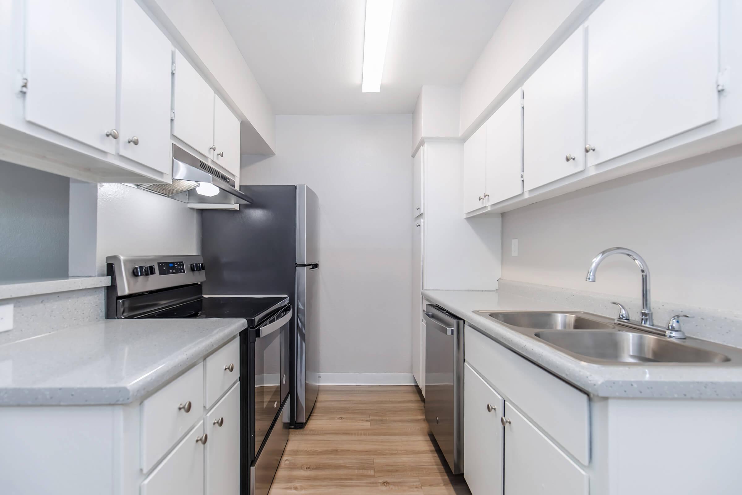 Modern kitchen with white cabinetry, stainless steel appliances including a refrigerator, oven, and dishwasher. The countertops are light-colored and the flooring is light wood. The space is well-lit with overhead lighting, creating a clean and inviting atmosphere.