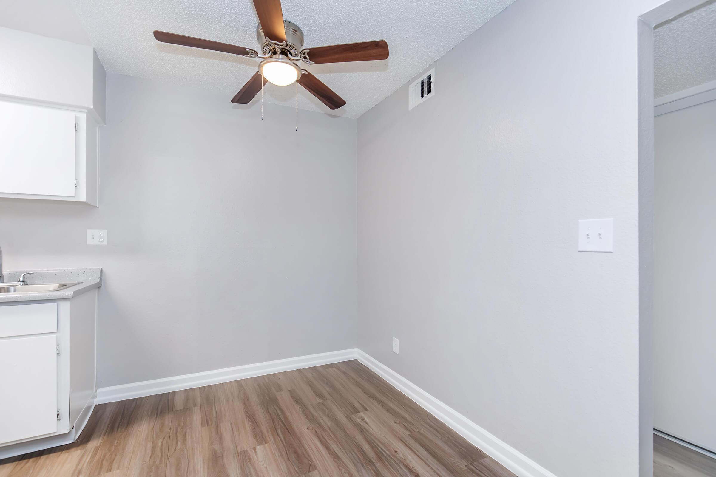 A simple, well-lit room featuring a ceiling fan with wooden blades, light gray walls, and laminate flooring. There is a small kitchen area with white cabinets and a sink visible to the left, while a doorway leads to another space out of view. The atmosphere is clean and modern.