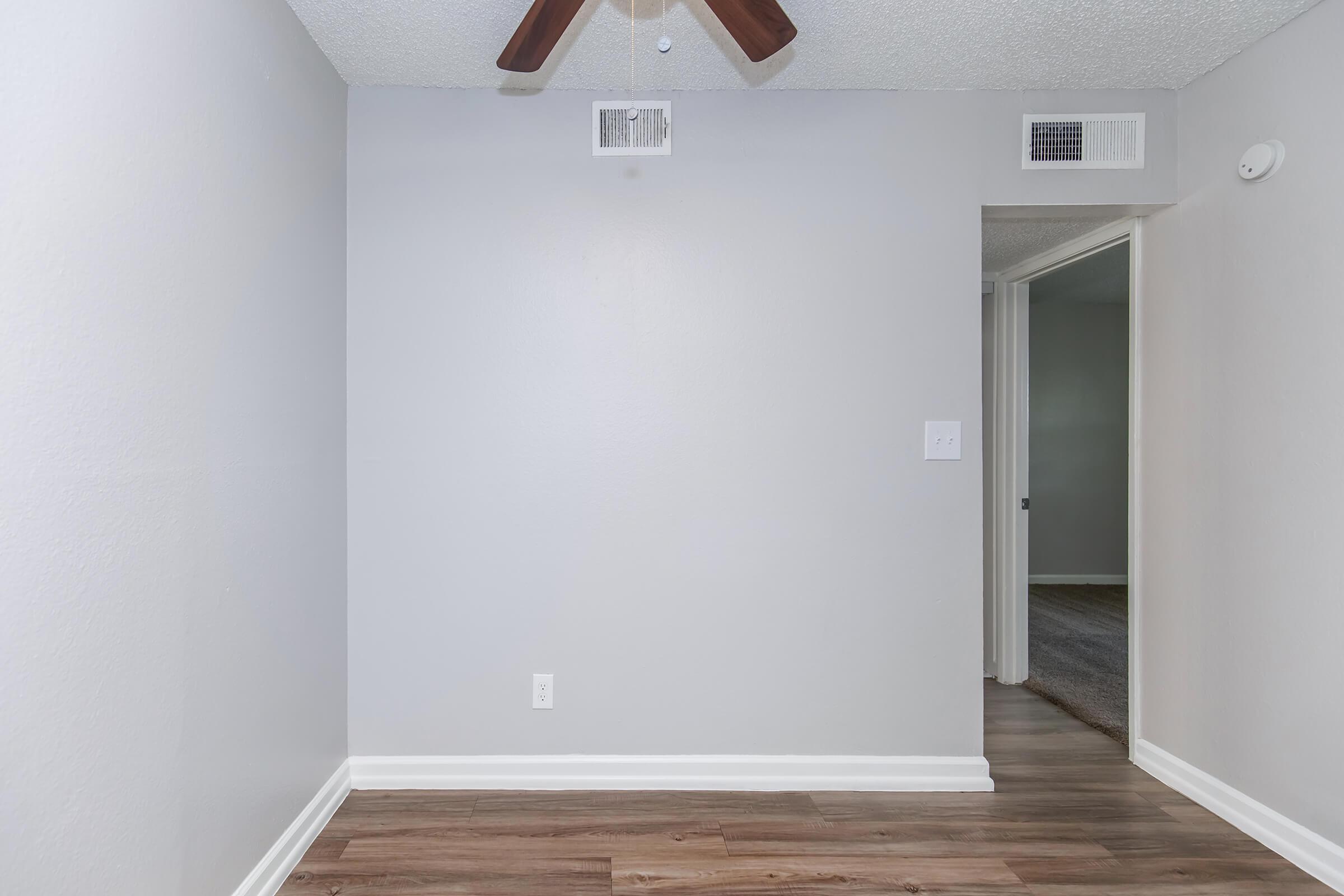 An empty room with light gray walls and a ceiling fan. The flooring is wooden, and there is a doorway leading to another room on the right. Air vents are visible on the walls, and there is a light switch on the left side. The overall atmosphere is simple and uncluttered.
