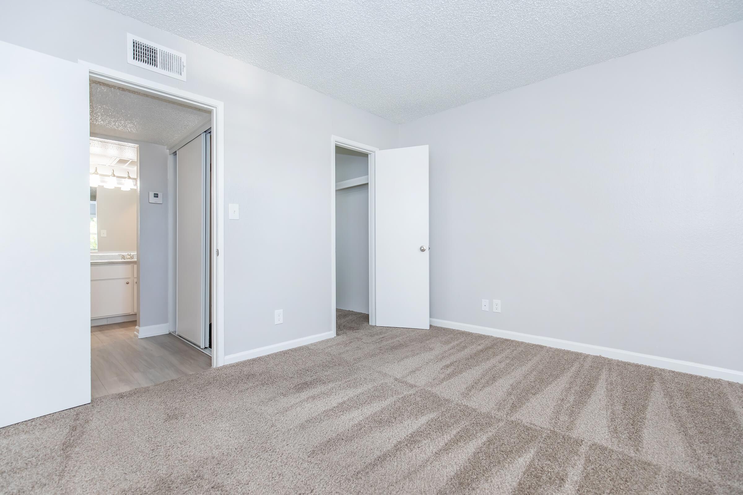 A spacious, well-lit room with light gray walls and plush carpet. There are two doorways leading to a bathroom and a closet. The entrance to the bathroom features modern lighting, enhancing the overall bright and inviting atmosphere of the space.
