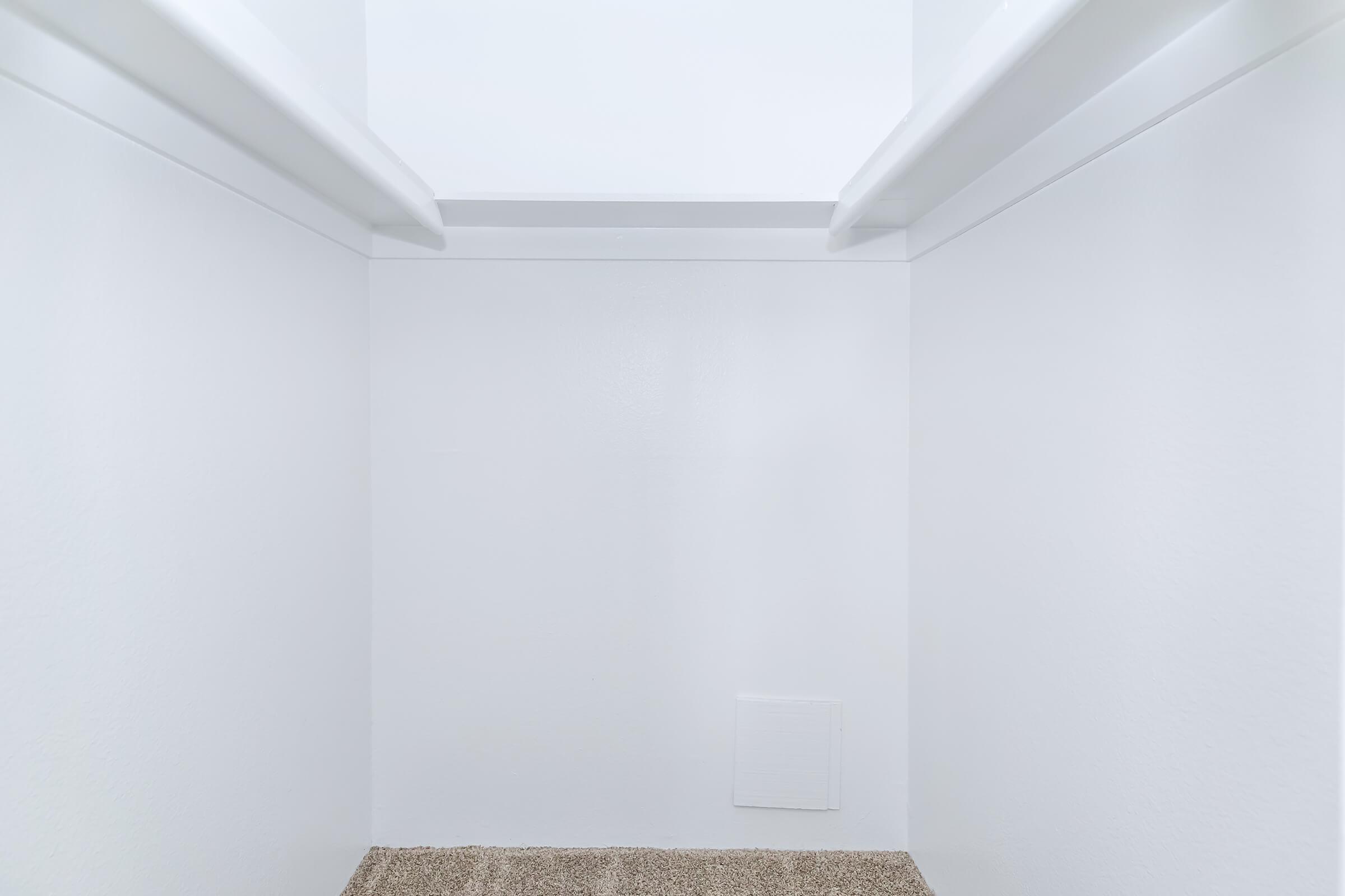 Empty closet interior with white walls, two shelves, and a beige carpeted floor. The backdrop is unobtrusive, featuring clear lines and a minimalistic design. There is a small vent or access panel on the lower right wall.