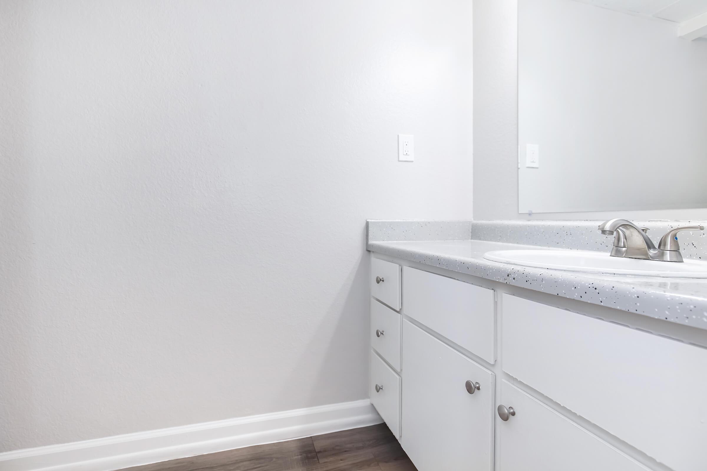 A clean and modern bathroom with a light gray wall. The countertop is light with speckles, featuring a sink and faucet. White cabinetry with knobs is visible beneath the countertop. A mirror reflects the simple design, enhancing the bright and airy ambiance of the space.