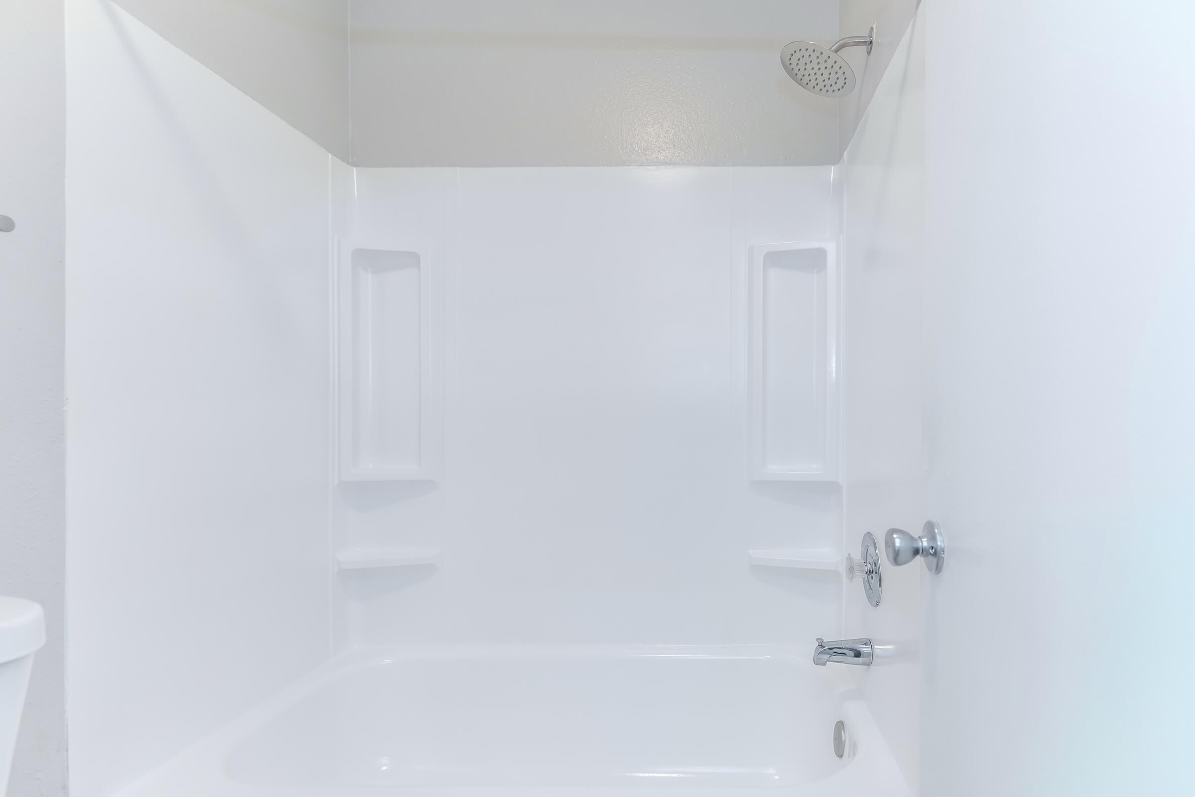 A clean, white bathtub and shower enclosure with a handheld showerhead and two built-in shelves for toiletries. The walls are smooth and the space is well-lit, giving a fresh and modern appearance.