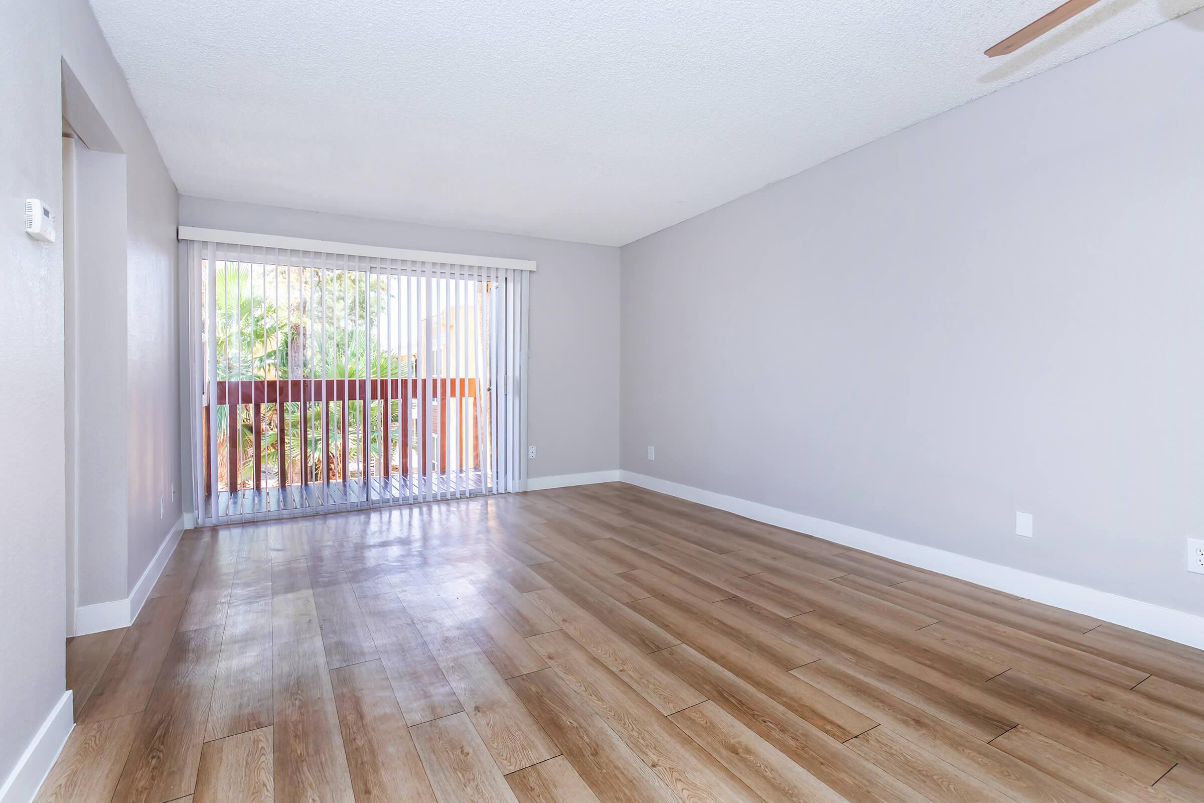 A spacious, well-lit living room featuring light wooden flooring and neutral-colored walls. Large sliding glass doors with vertical blinds lead to a balcony with greenery outside. The room has a ceiling fan and an unobstructed view, creating a bright and airy atmosphere.