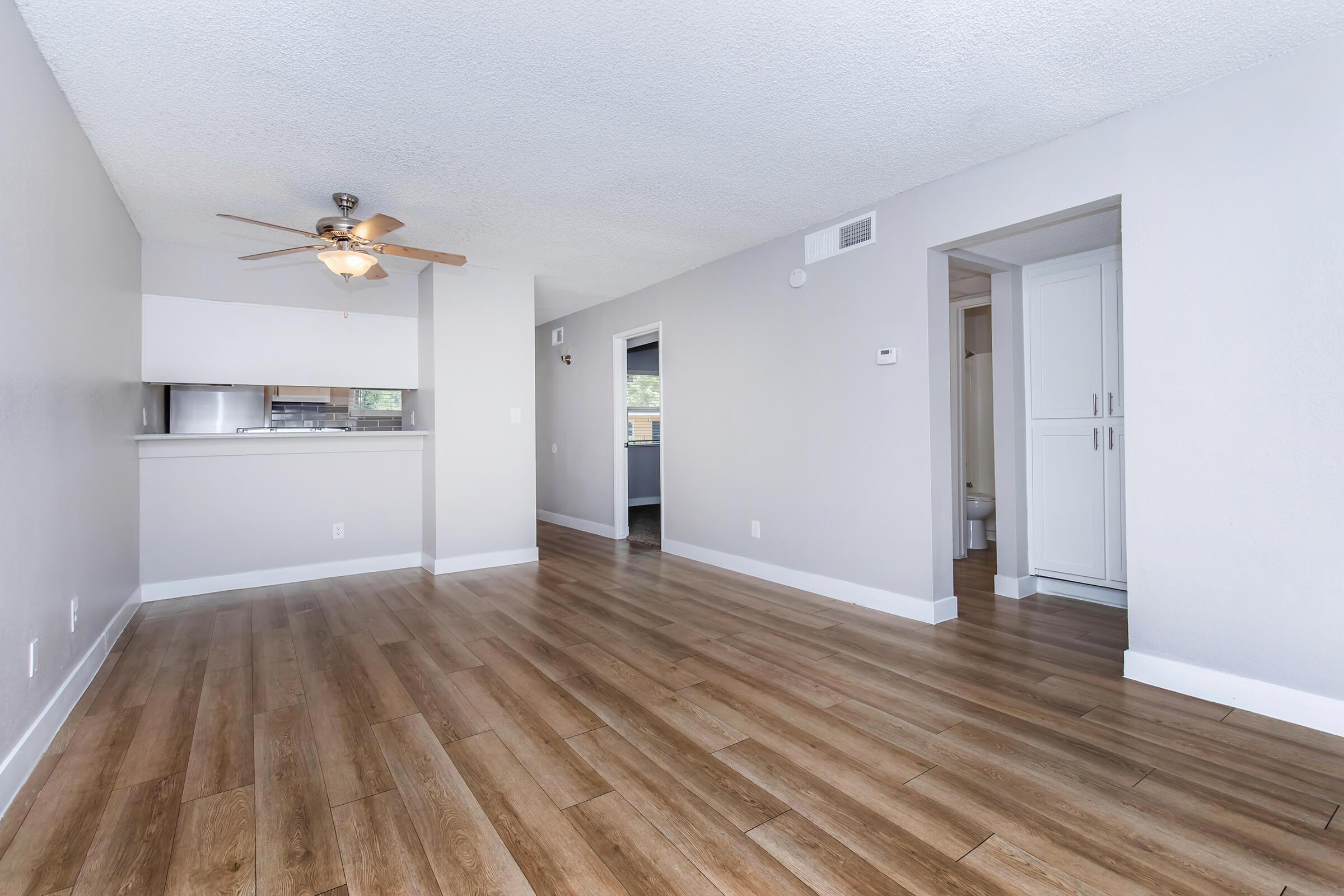 Spacious living area with wood-like flooring, a ceiling fan, and neutral-colored walls. The layout includes a doorway leading to another room and an open kitchen with a breakfast bar visible in the background. Natural light brightens the space, creating a welcoming atmosphere.