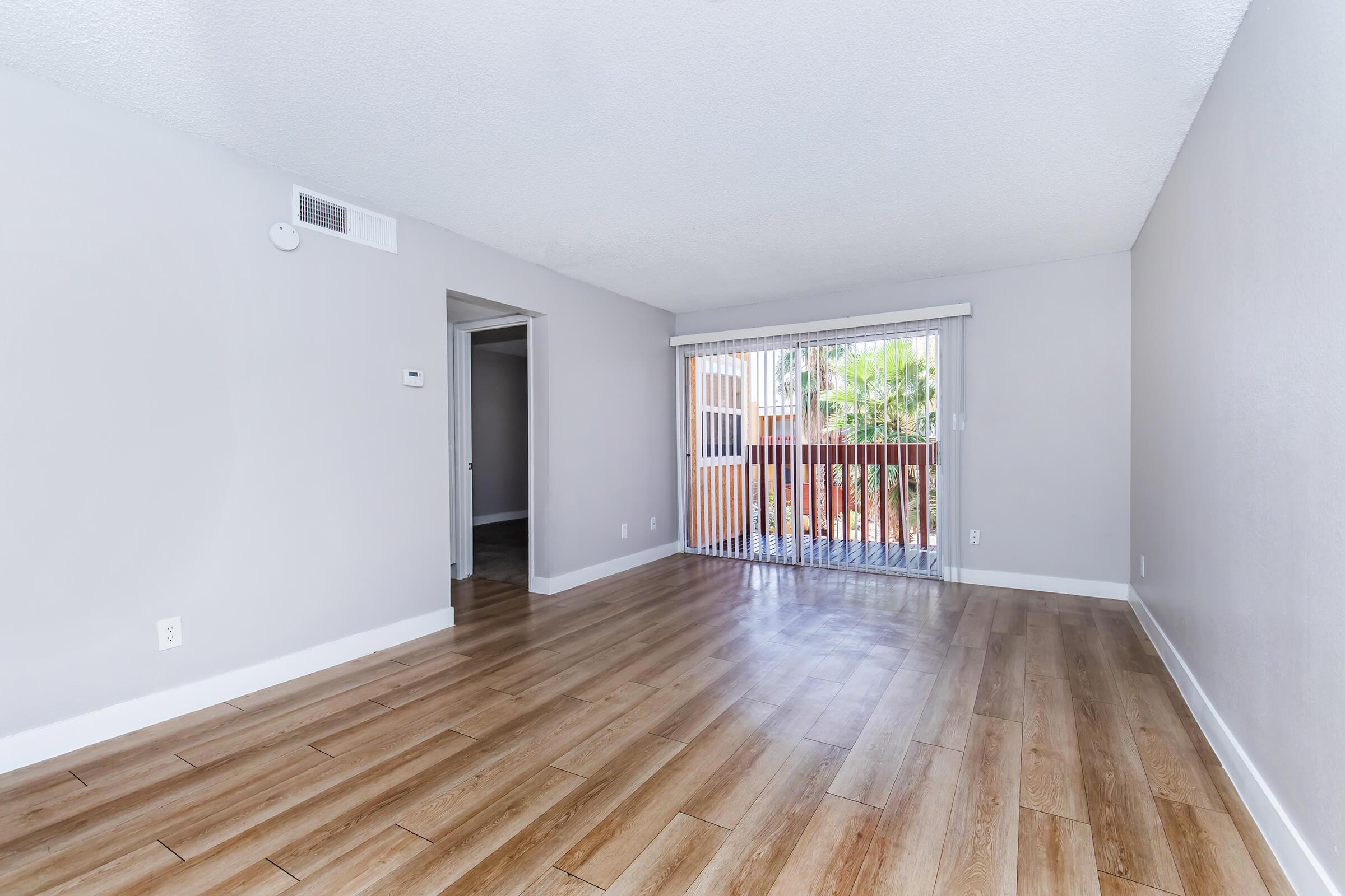 Spacious living room featuring light gray walls, wood-like flooring, and a sliding glass door leading to a balcony with outdoor views. The room is brightly lit and has a minimalist design, with a small entryway visible on the left.
