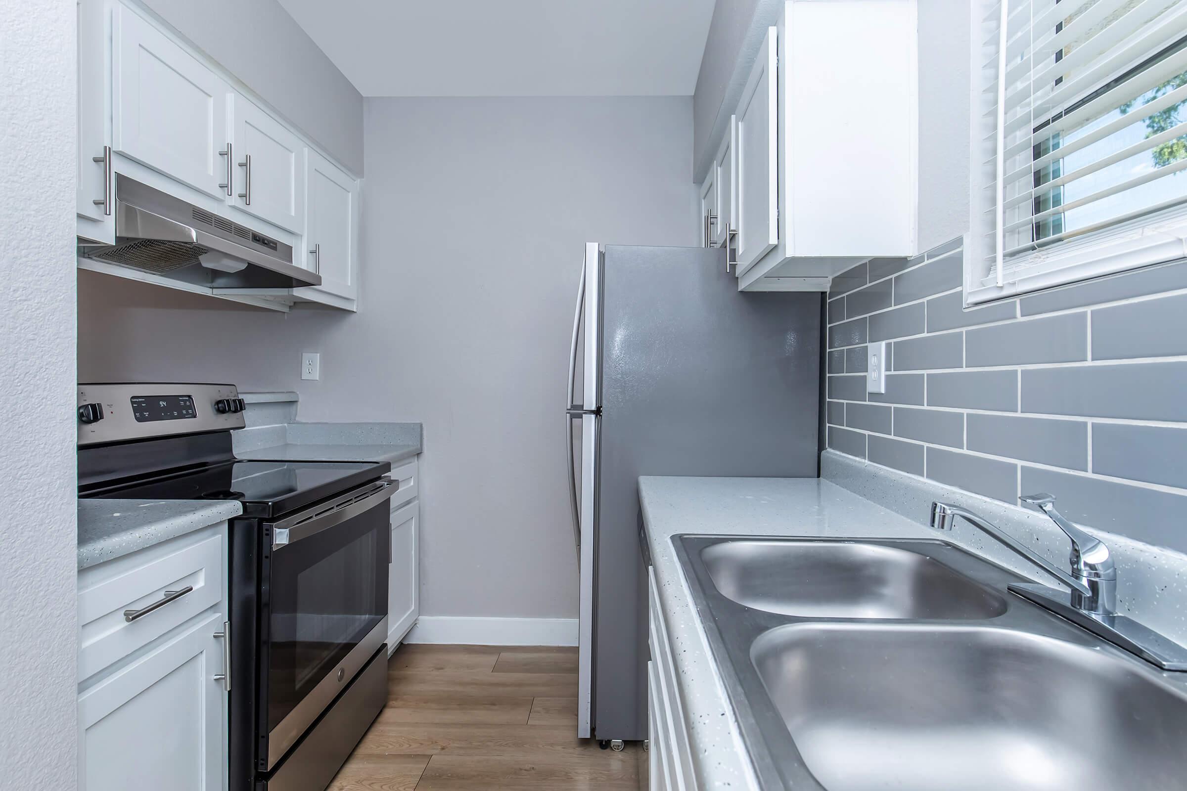 A modern kitchen featuring stainless steel appliances, including an oven and refrigerator. The cabinetry is white, with light gray walls and a stylish backsplash. A double sink is positioned beside the window, allowing natural light into the space, which has a warm wooden floor.
