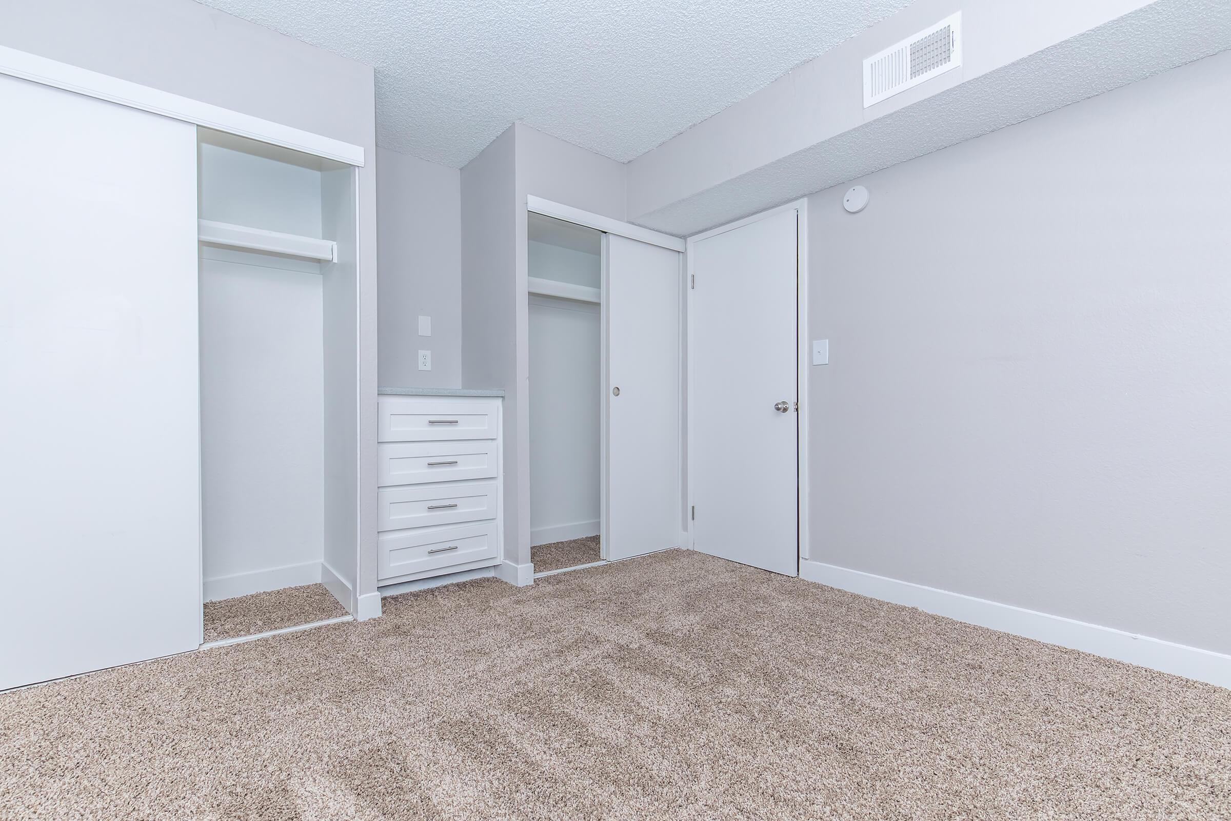 Empty bedroom featuring light gray walls and carpet. The room includes a closet with sliding doors on the left, a set of drawers, and a closed door on the right. Natural light brightens the space, creating a clean and modern atmosphere.