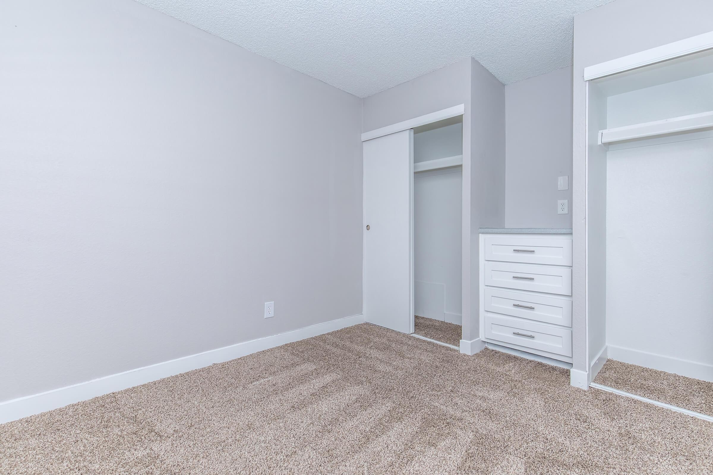 A simple, empty room with light gray walls and plush beige carpet. There's a built-in closet with sliding doors on the left, and to the right, a chest of drawers with three white drawers. The room has a clean, minimalistic design, with a bright, airy feel.