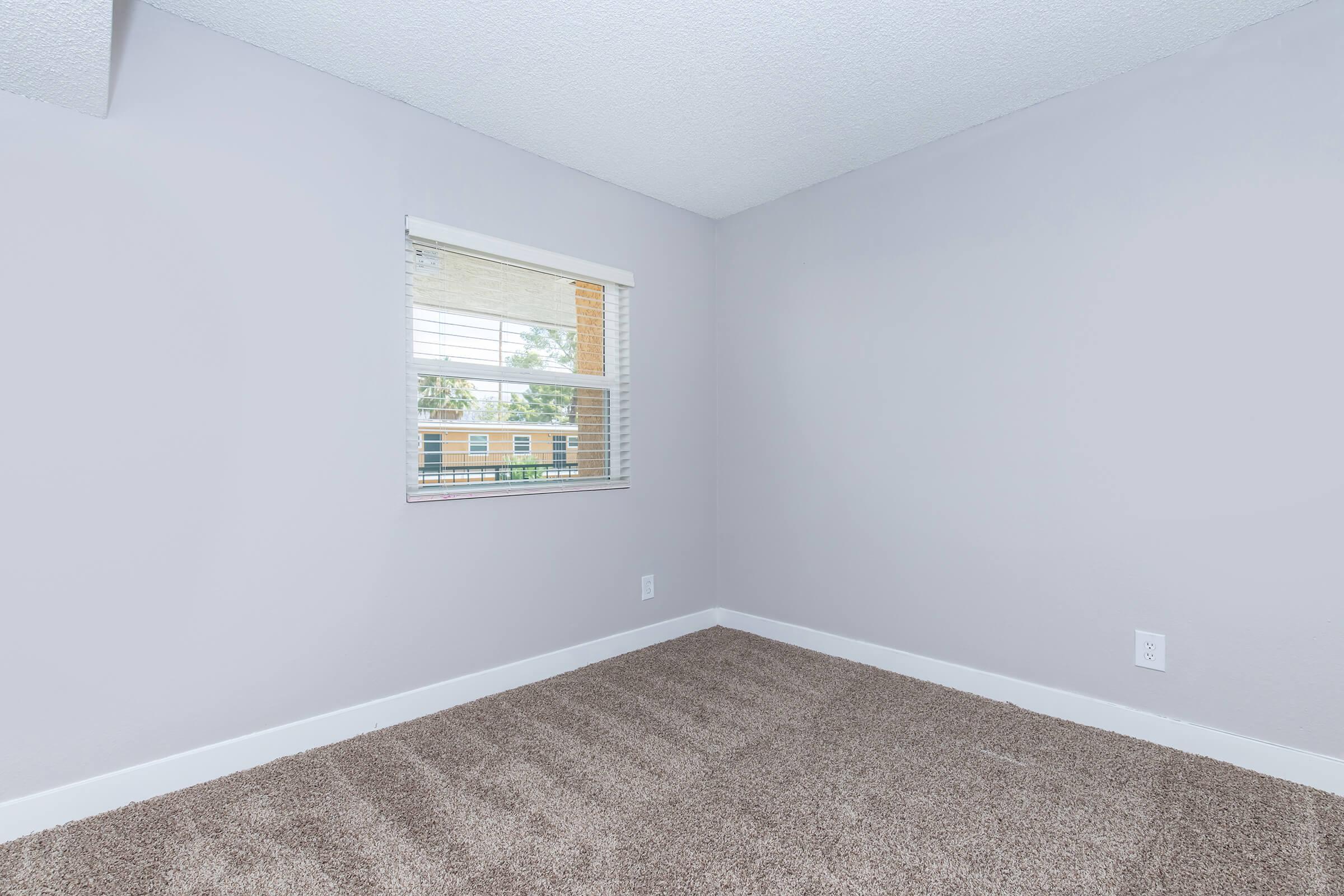 A small, vacant room with light gray walls and beige carpeting. There is a window with white blinds allowing natural light to enter, and a subtle hint of greenery visible outside. The space is simple and uncluttered, suggesting potential for various uses.