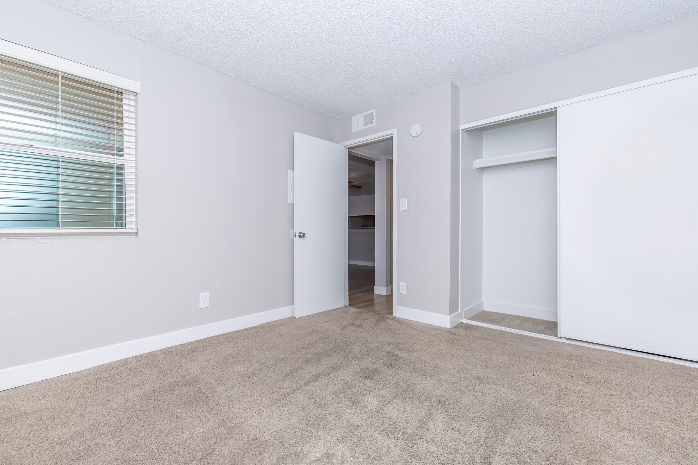 A small, carpeted bedroom with light gray walls. There is a window with blinds on the left side, and a white door leading to another room is slightly ajar. A closet with sliding doors is visible on the right side, and the room is devoid of furniture. The lighting is bright and neutral.