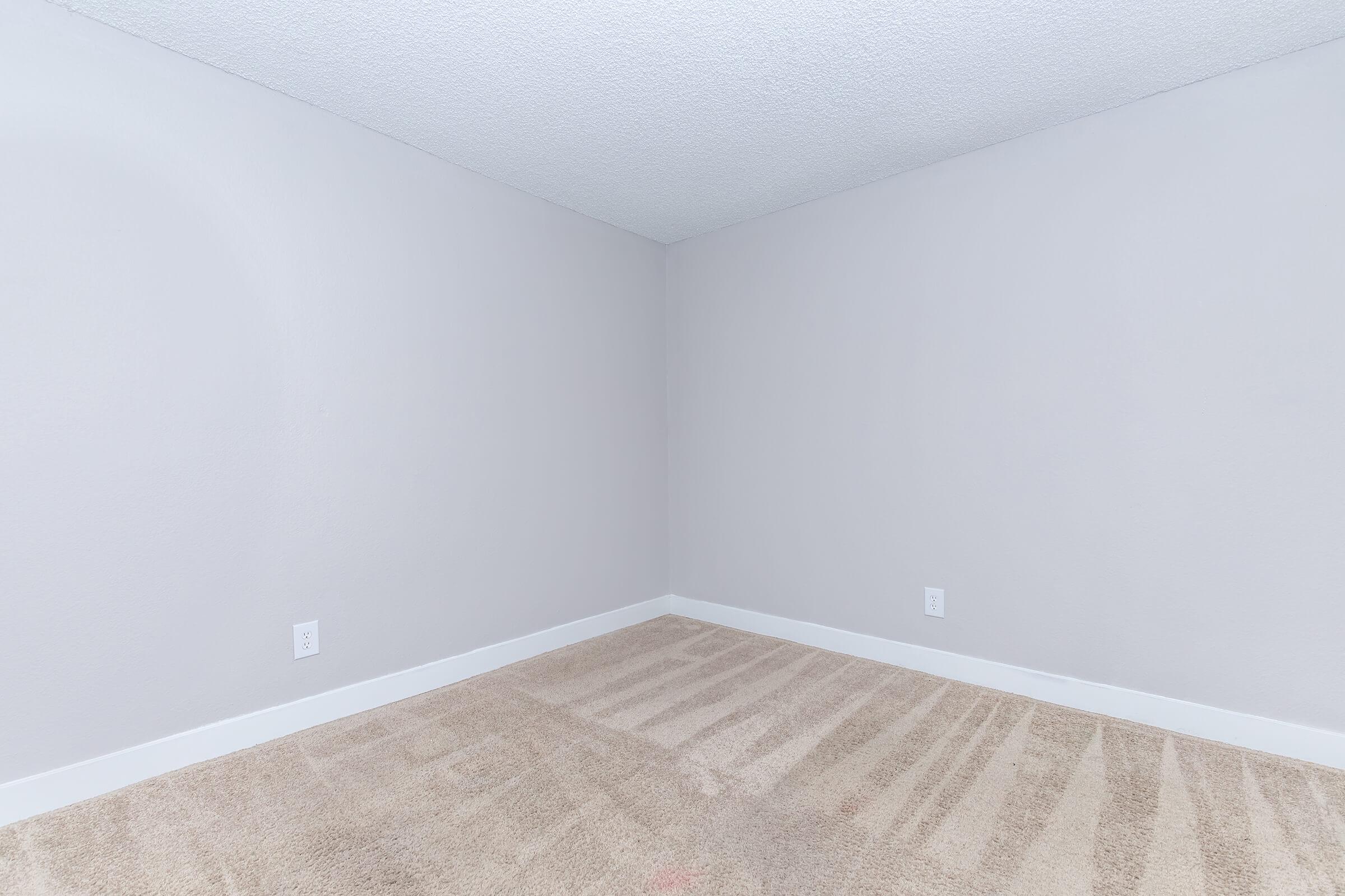 A vacant room with light gray walls and beige carpet. The room features a corner with no furniture or decor, emphasizing a simple, minimalistic design. Two electrical outlets are visible on the walls, and the ceiling has a textured finish. Natural light may fill the space, creating a bright atmosphere.