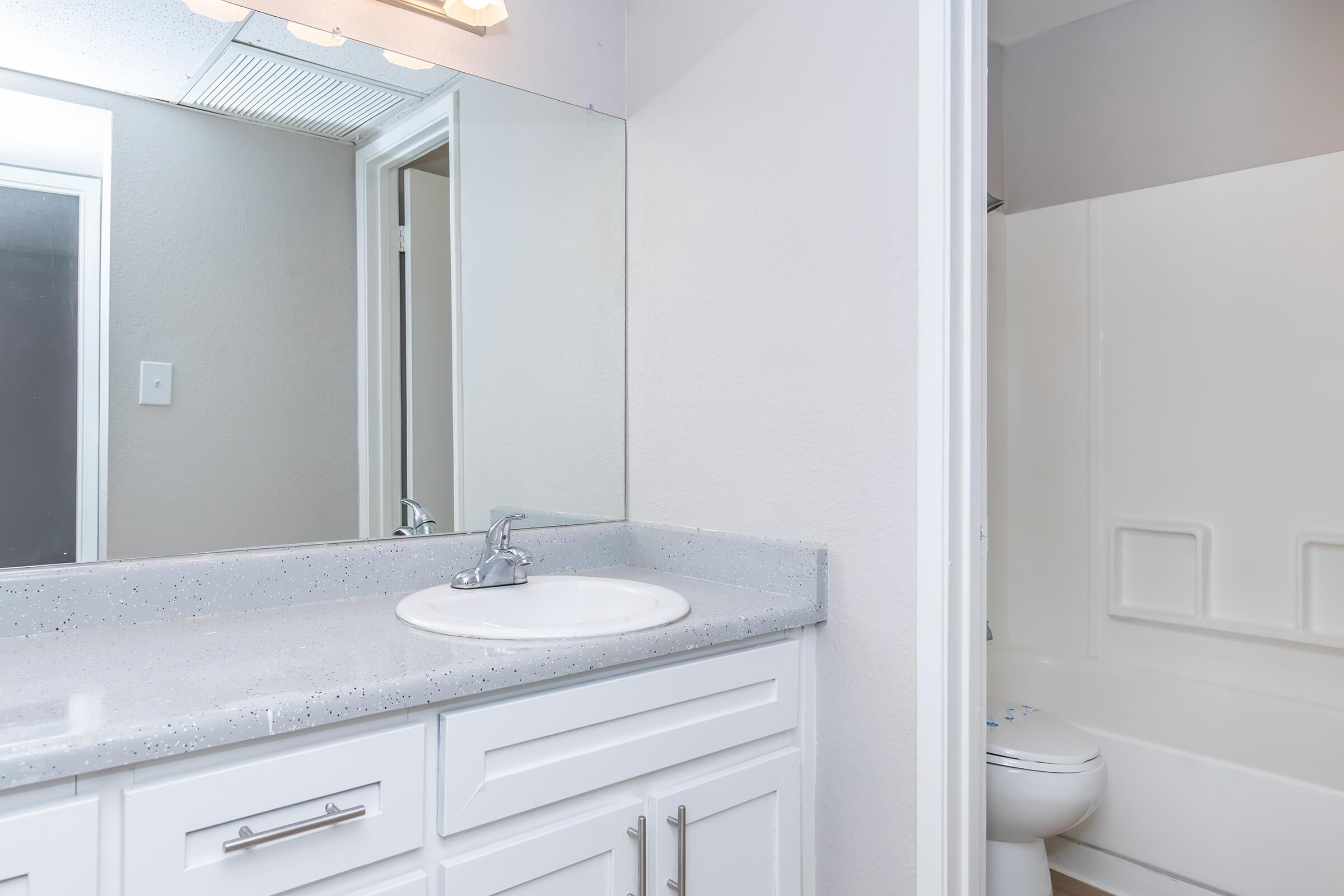 Modern bathroom featuring a sleek white vanity with a round sink, a light gray countertop, and a large mirror above. The space includes a bath/shower combo on the right and a door leading to another area, with neutral-colored walls enhancing the bright, clean look.
