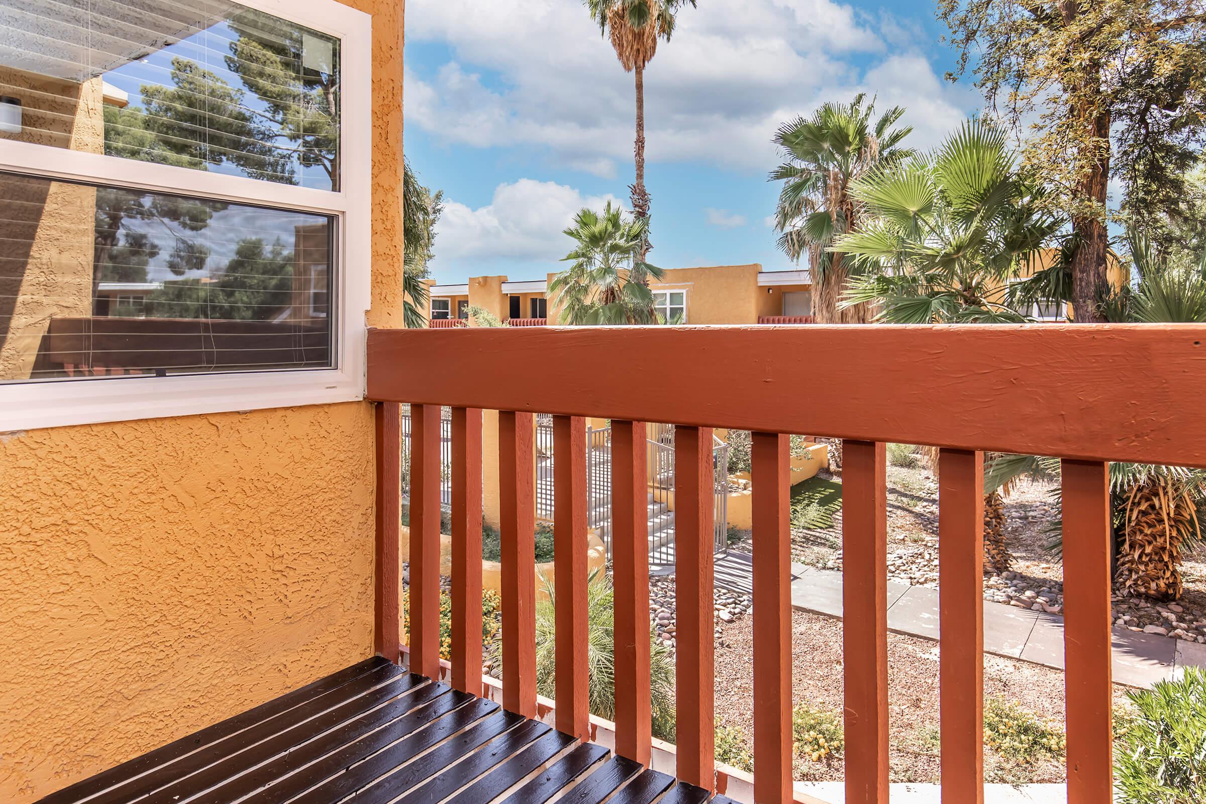 View from a balcony showing a wooden railing and painted wall, with palm trees and colorful buildings in the background against a partly cloudy sky. The scene captures a relaxed outdoor atmosphere in a landscaped environment.
