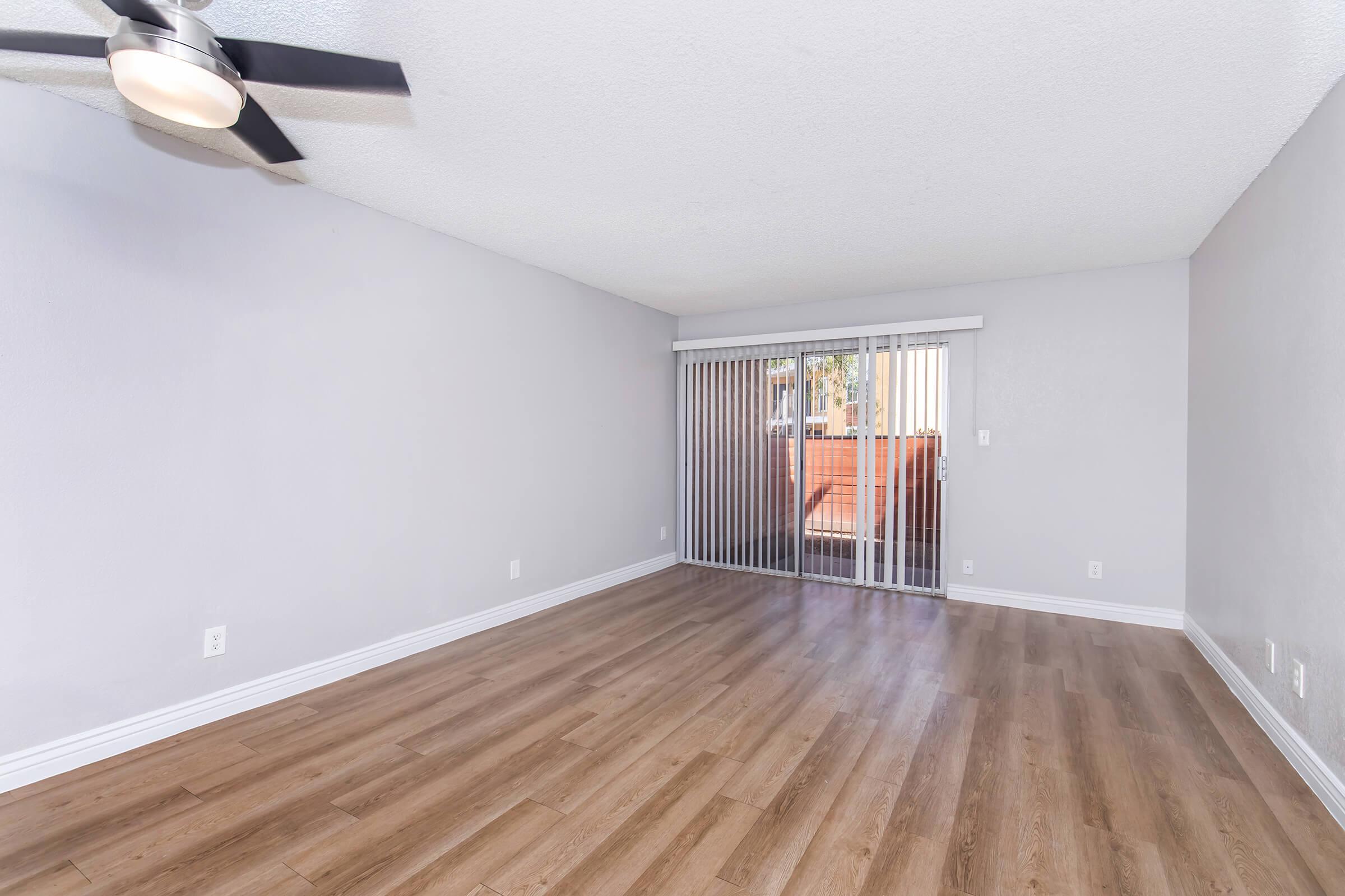 A spacious, empty living room featuring light gray walls, a ceiling fan, and large windows covered with vertical blinds. The floor is covered in wooden laminate, creating a warm atmosphere. A balcony is visible through the sliding doors, allowing natural light to enter the space.