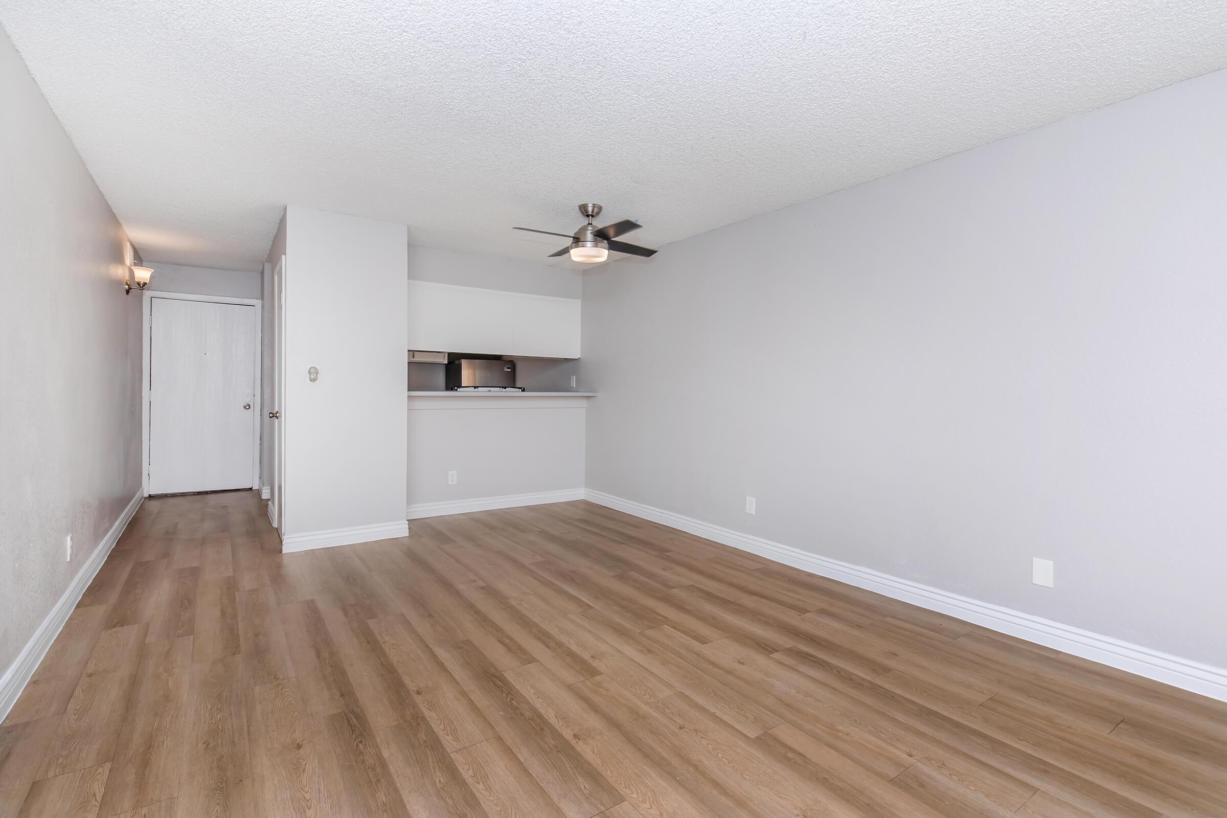 A spacious, empty living room featuring light gray walls, a ceiling fan, and wood laminate flooring. The room is well-lit with natural light, and there is a doorway leading to another room. A small kitchen area is visible in the background, partially separated by a counter.