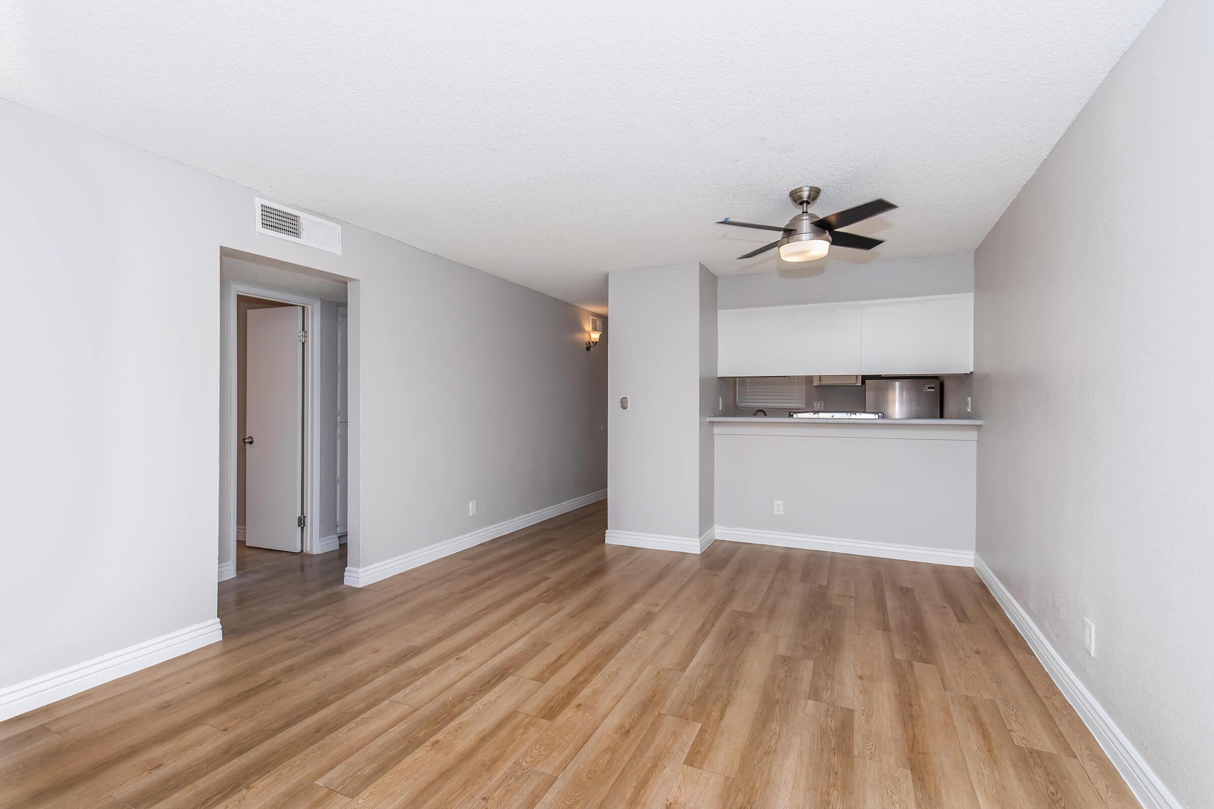 A spacious, modern living area with light gray walls and new wood-like flooring. A ceiling fan is visible, and there's an open layout leading to a kitchen area. A doorway can be seen on the left, suggesting access to another room. The space is bright and inviting.