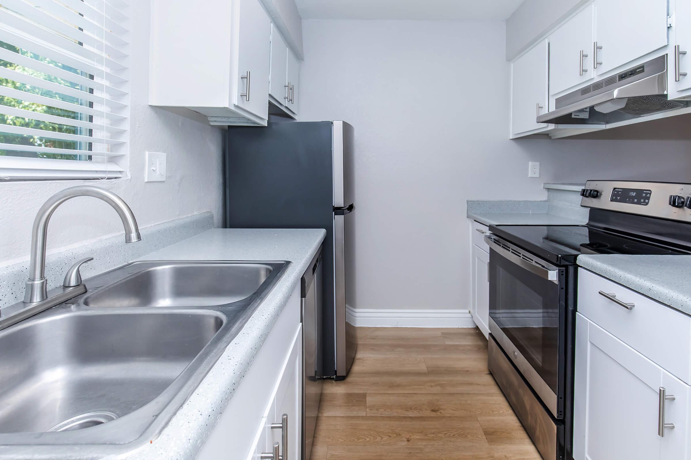 Modern kitchen featuring stainless steel appliances, including a refrigerator and oven. The countertops are light-colored with a double sink, and there are white cabinets. Natural light enters through a window with blinds, illuminating the wooden floor.