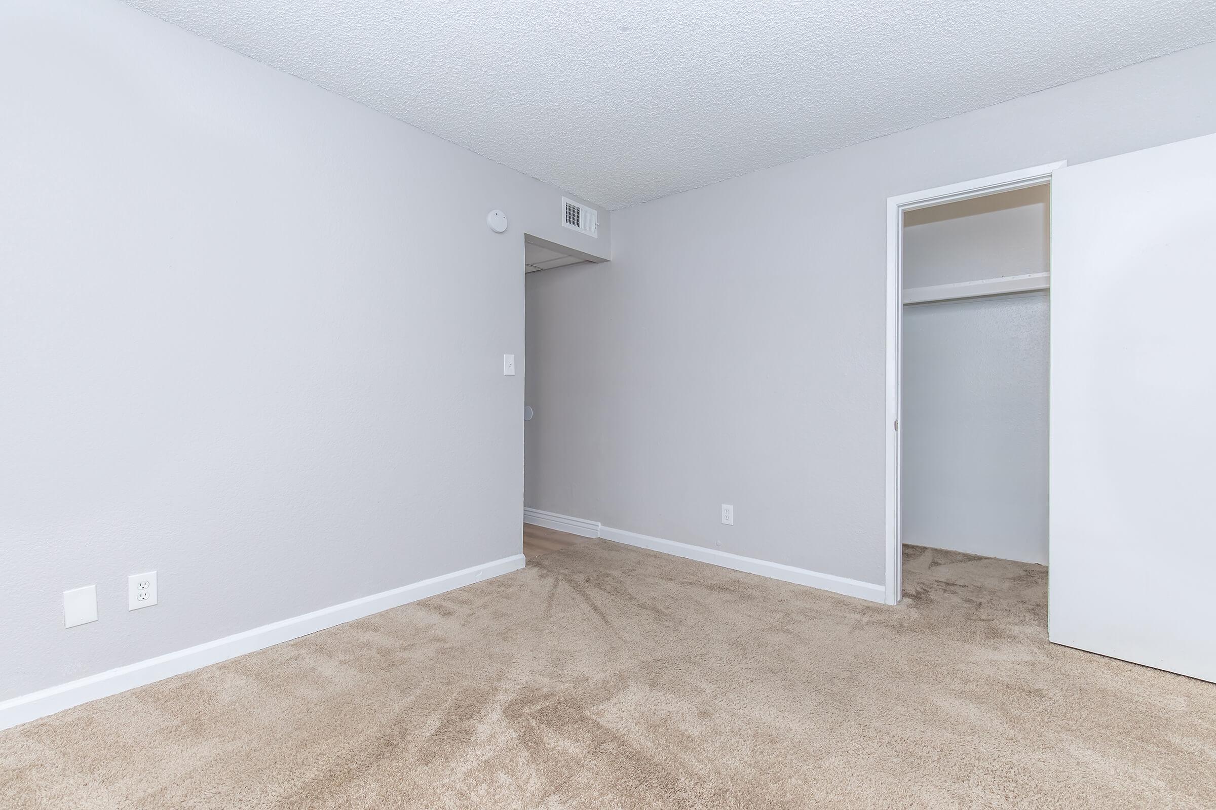 Empty room with light gray walls and beige carpet. A closet door is partially visible on the right, and there's an open doorway leading to another space. The room has no furniture or decorations, creating a minimalist and spacious appearance. Natural light fills the area, enhancing the neutral colors.