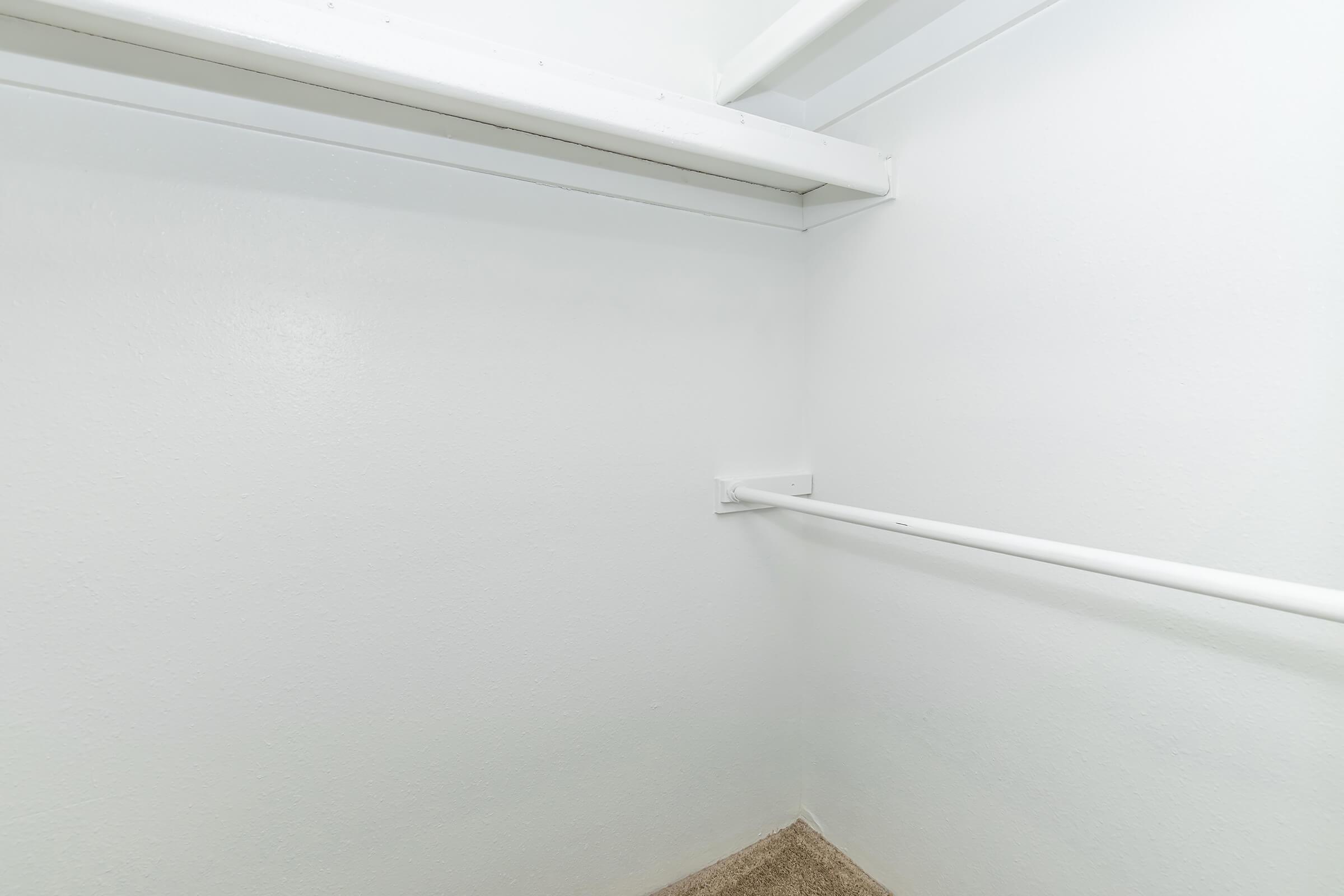 Empty closet with white walls and a single horizontal clothing rod. The floor is carpeted in a neutral color. The space is well-lit, giving it a clean and minimalist appearance.
