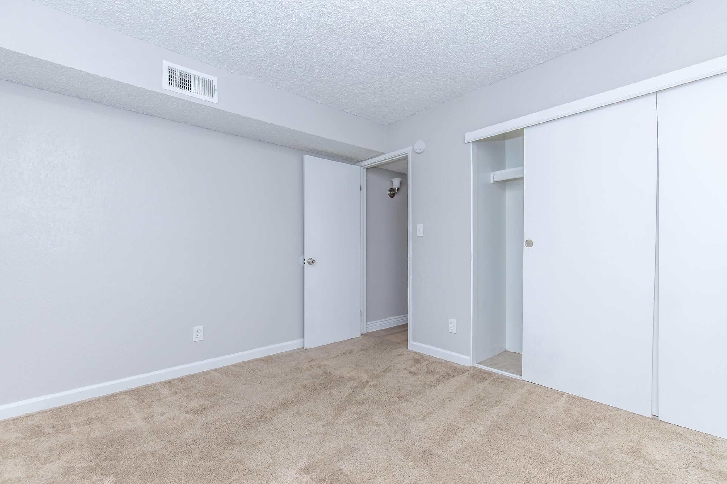 A clean and empty room with light gray walls and a carpeted floor. There are two closed doors: one leading to a closet and another to the hallway. Natural light illuminates the space, which has no furniture, creating a minimalist and open atmosphere.