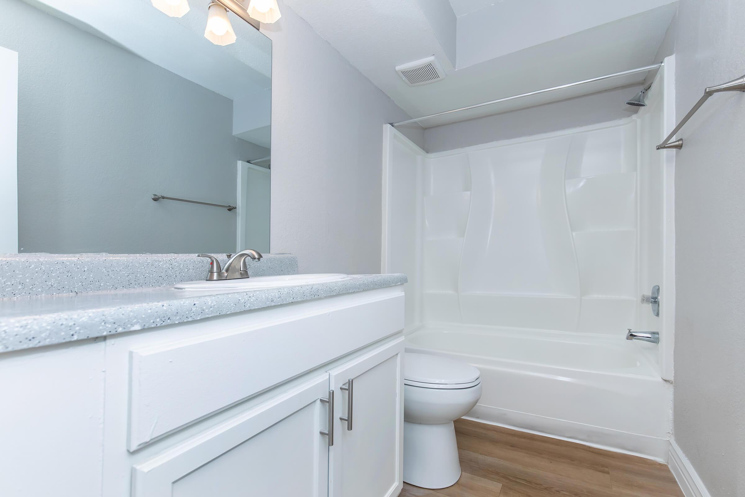 Bright and clean bathroom featuring a white countertop with a sink, a bathtub with a shower, and a modern toilet. The walls are painted in a soft gray, and the space is well-lit with overhead lighting. The floor has a wooden finish, adding warmth to the contemporary design.
