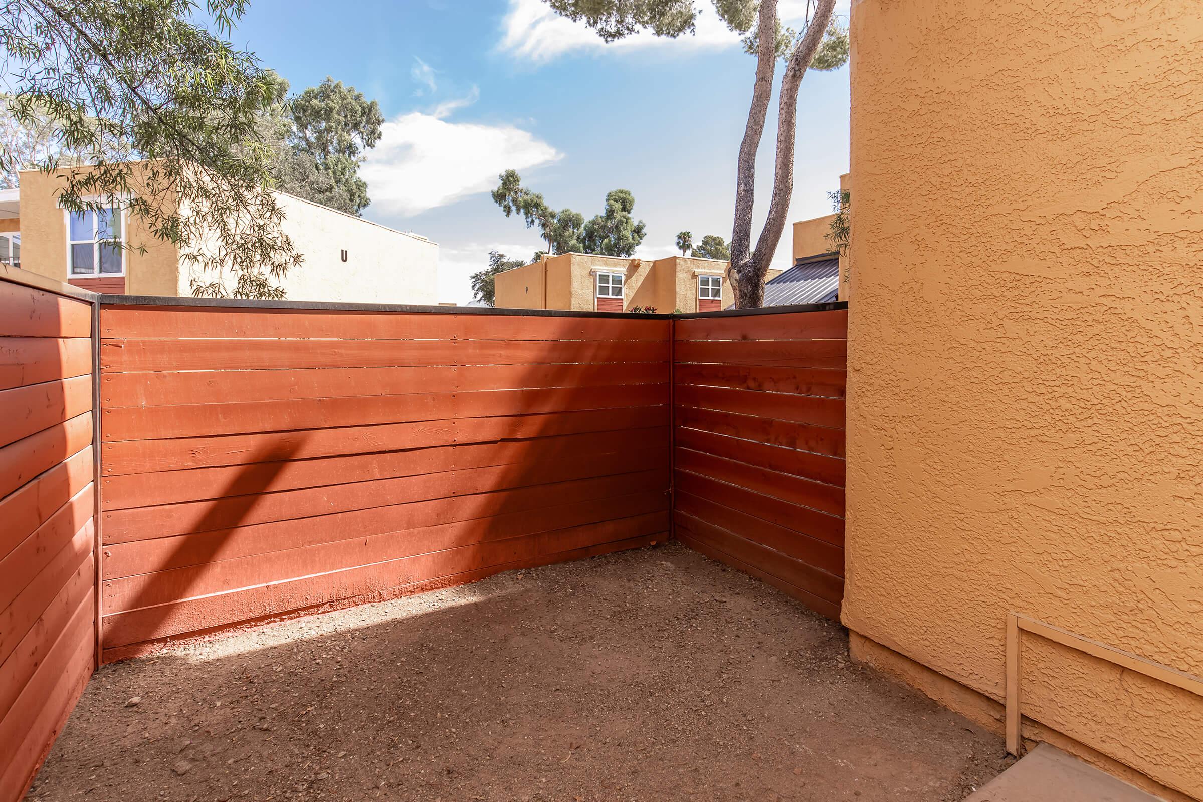 A corner of a yard enclosed by a brown wooden fence, with a dirt ground. In the background, there are buildings with windows, and trees are visible against a blue sky with some clouds.