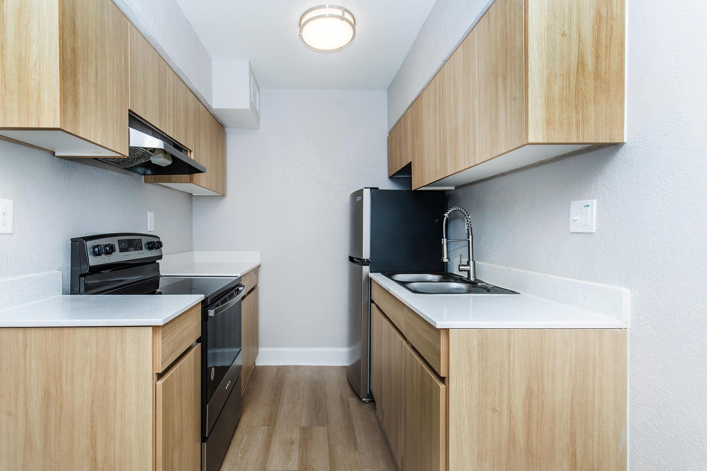 Modern kitchen with light wood cabinets, a black stove and refrigerator, and a stainless steel sink. The countertops are white and the flooring is light-colored wood. The walls are painted a soft gray, and there is a round ceiling light fixture.