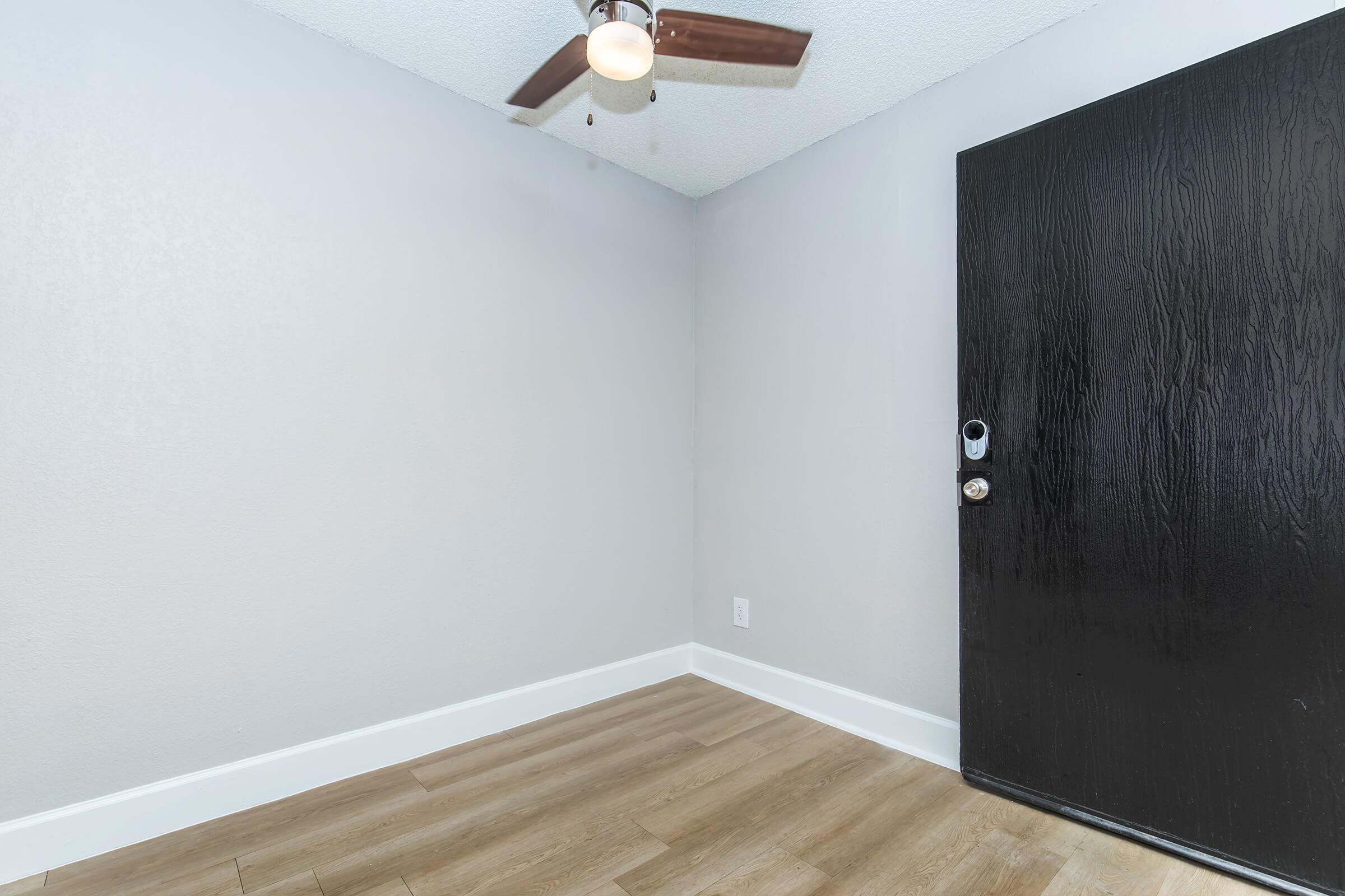 A small room with gray walls and a ceiling fan. There is a dark front door on the right side and light-colored flooring. The space appears empty and well-lit, creating a simple and modern aesthetic.
