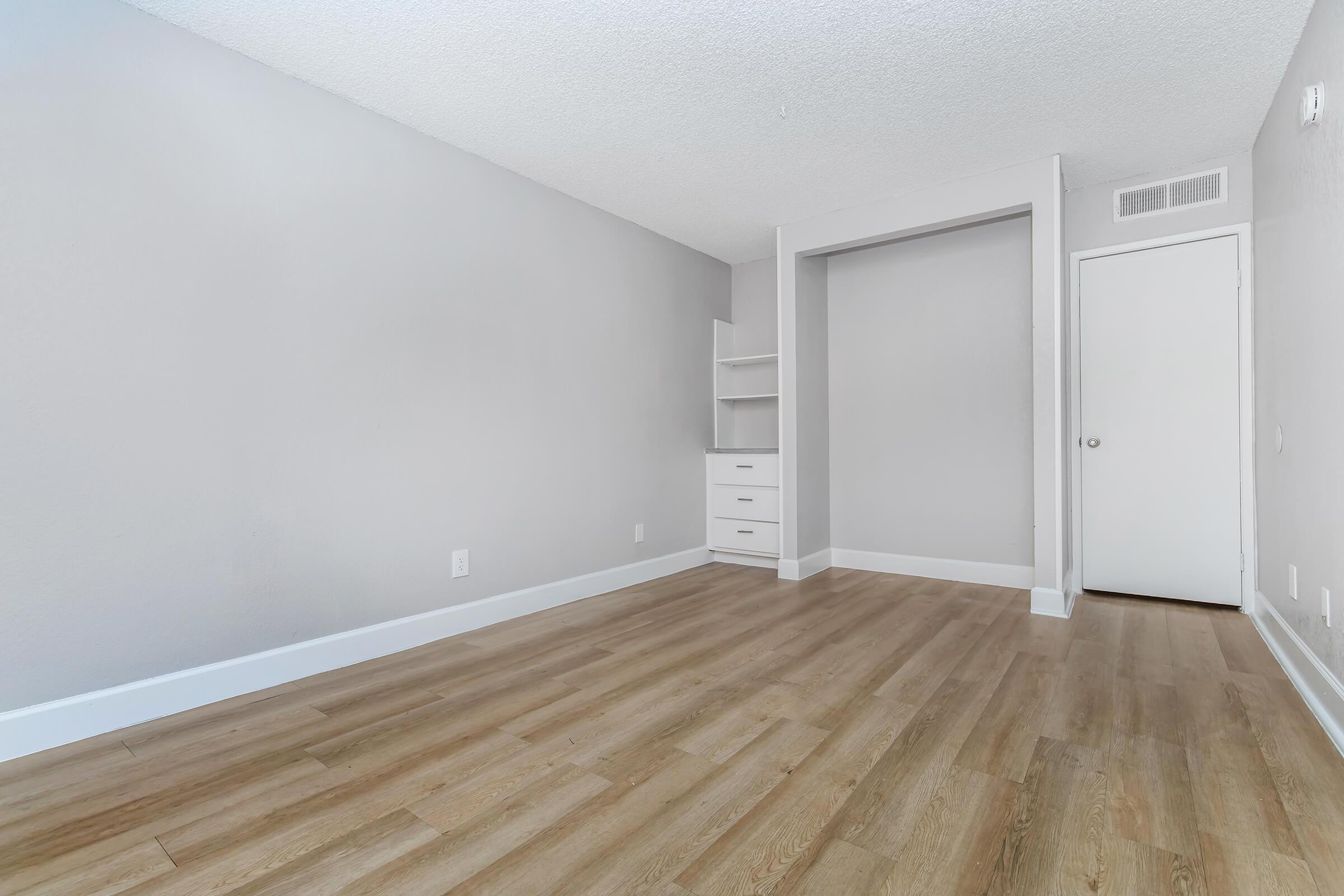 A spacious, empty room with light gray walls and hardwood flooring. It features a closet nook, a built-in shelf, and a white door. The overall atmosphere is bright and clean, suitable for various purposes such as a bedroom or office.
