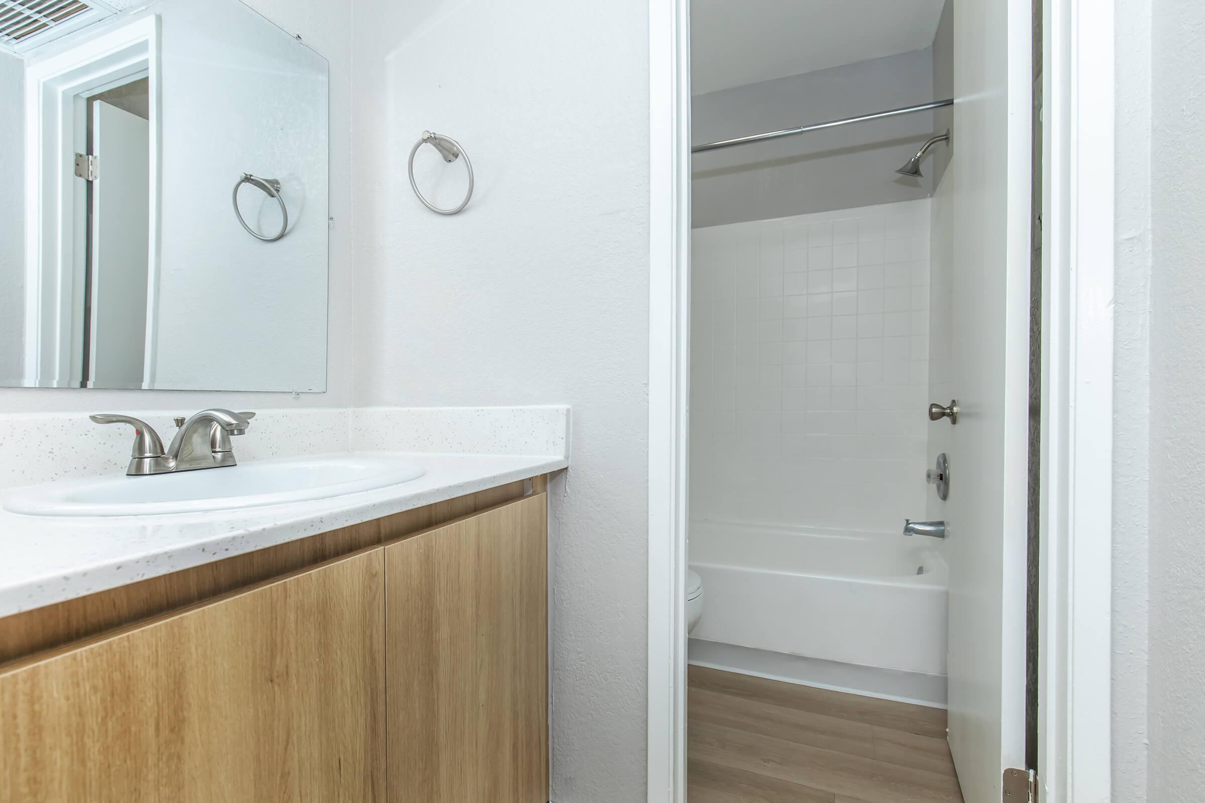 A modern bathroom featuring a sink with a mirror above, a wooden cabinet below, and a shower/tub combination visible in the background. The walls are painted white, and the floor has a light wood finish, creating a clean and bright atmosphere.