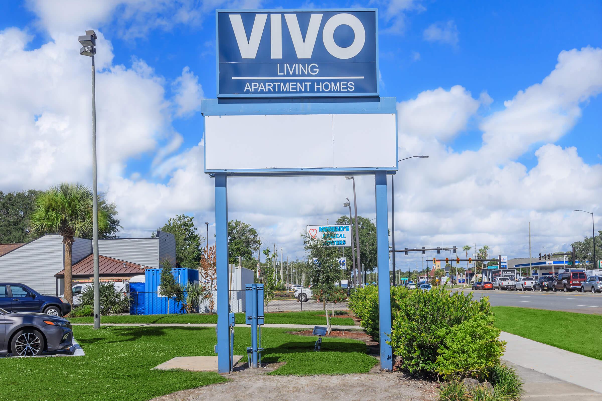 Sign for Vivo Living Apartment Homes, situated outside with a clear blue sky and scattered clouds in the background. The area is landscaped with greenery. A road and vehicles are visible in the distance, indicating a suburban setting.