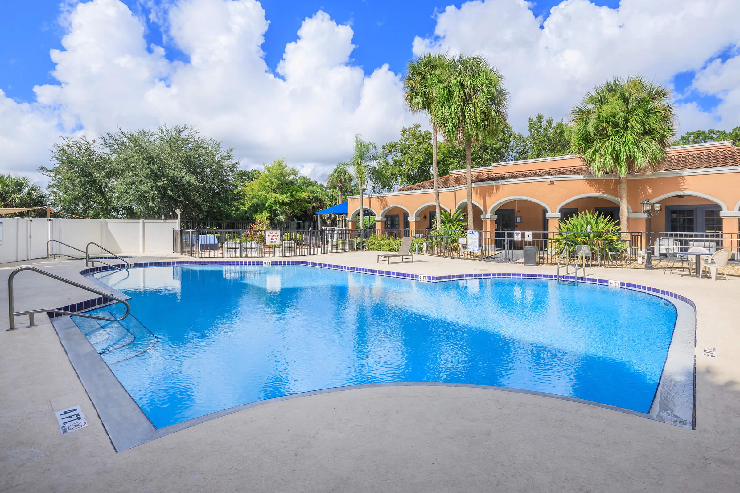 A clear blue swimming pool surrounded by a spacious deck, palm trees, and lush greenery. The area features lounge chairs and is bordered by a building with arches. The sky is bright with fluffy white clouds.