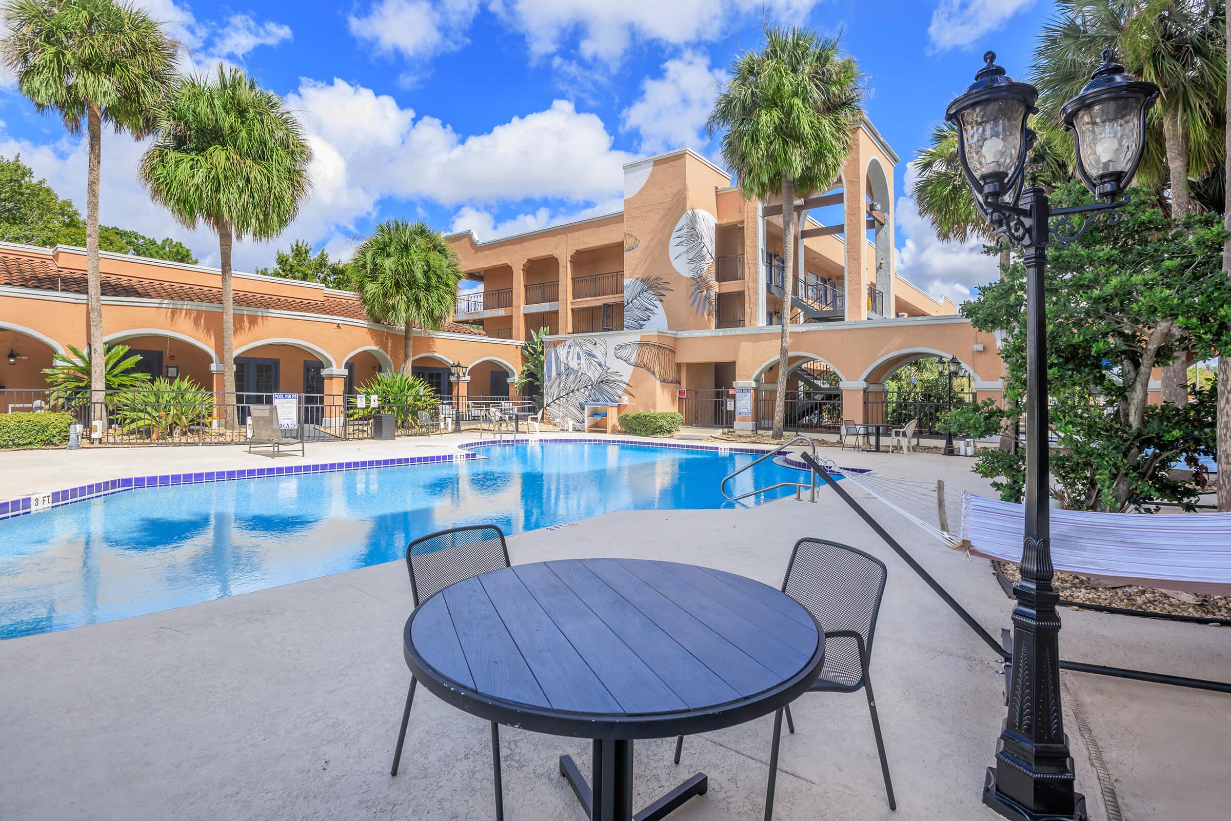 A serene pool area surrounded by palms and greenery, featuring a blue swimming pool and a table with chairs. In the background, there is a building with a mural and balconies, set against a bright sky with fluffy clouds. Lantern-style lighting adds charm to the outdoor space.