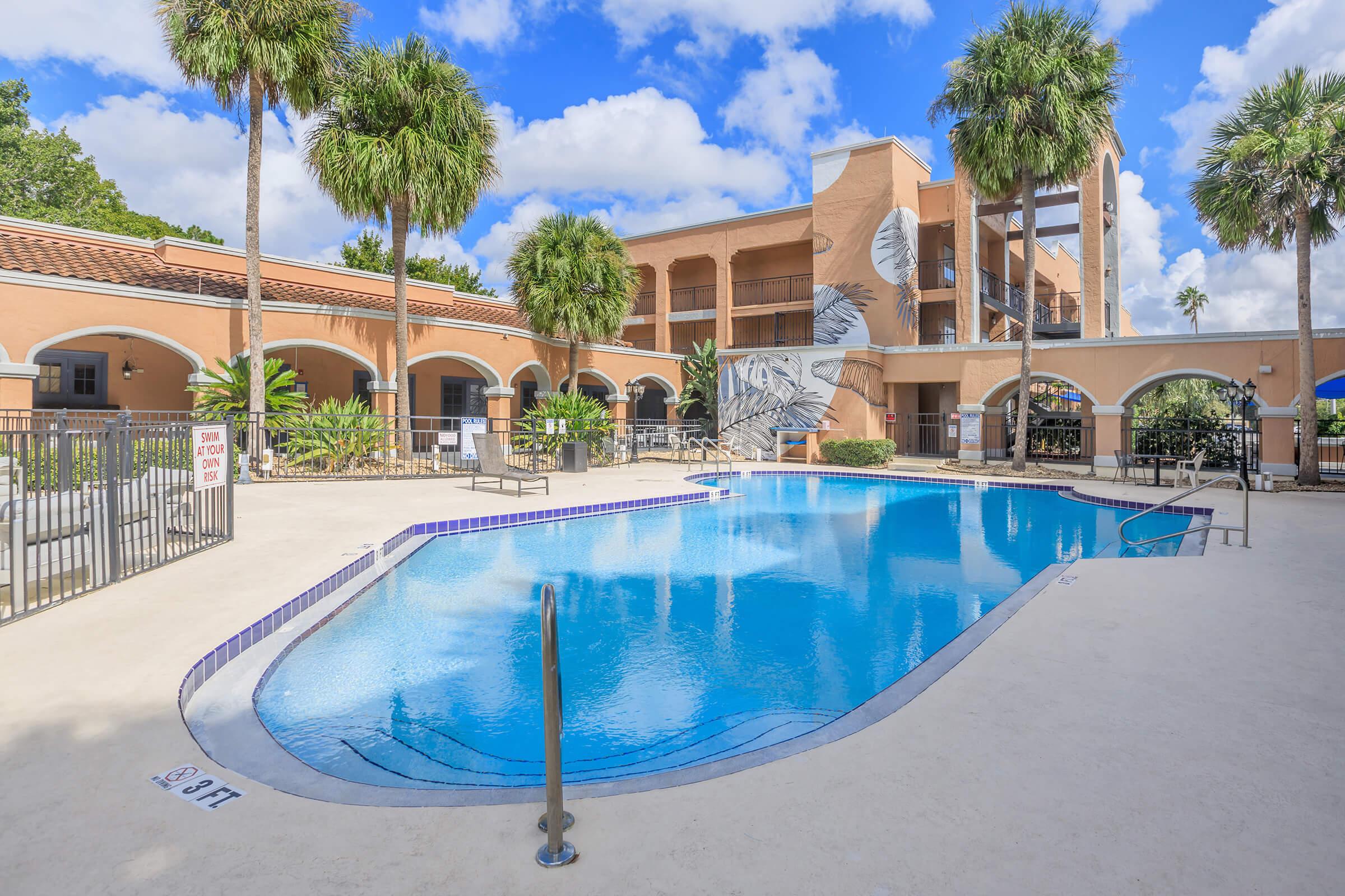 A clear blue swimming pool surrounded by palm trees and a spacious deck. The pool area features lounge chairs and is adjacent to a multi-story building with decorative elements. Lush greenery and a partly cloudy sky complete the pleasant outdoor setting.
