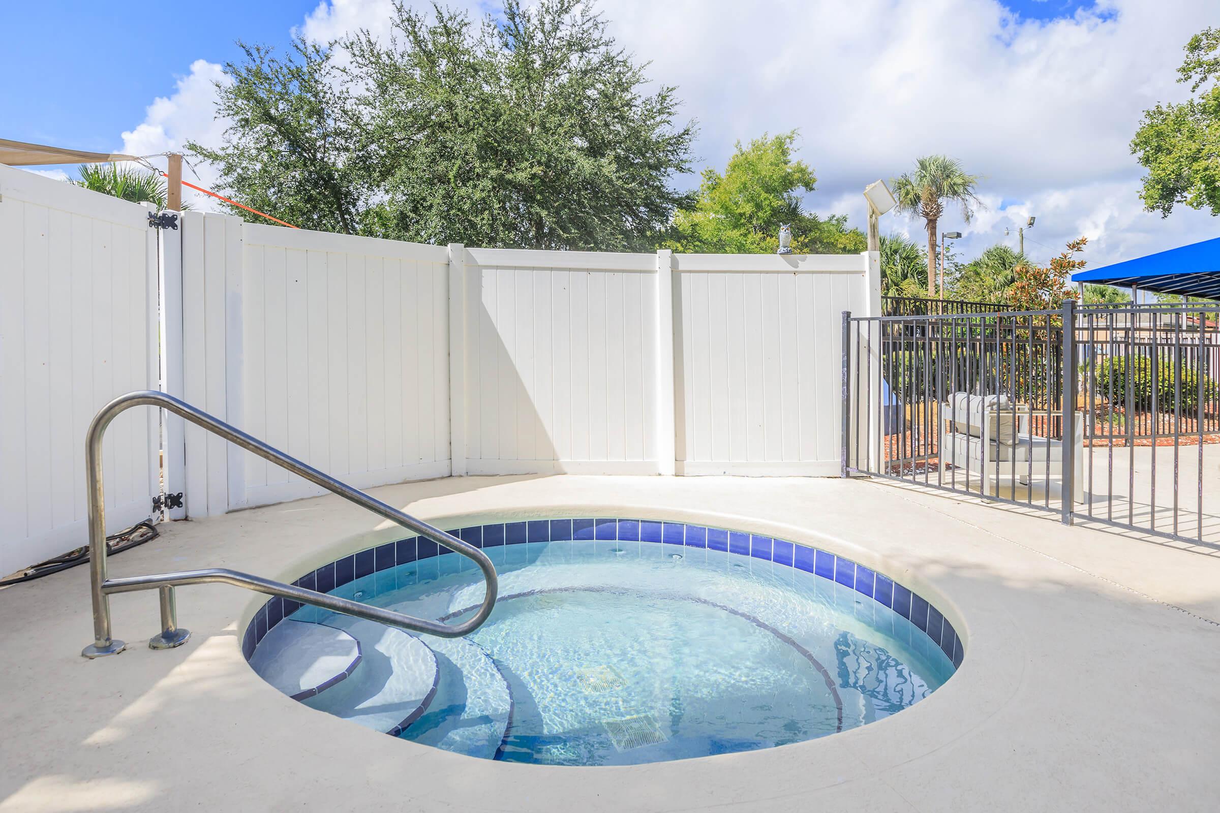 A round hot tub with a stainless steel railing, surrounded by a white fence. The area features green trees and blue skies with fluffy clouds in the background. A safety gate is visible nearby, and poolside furniture can be seen in the distance.