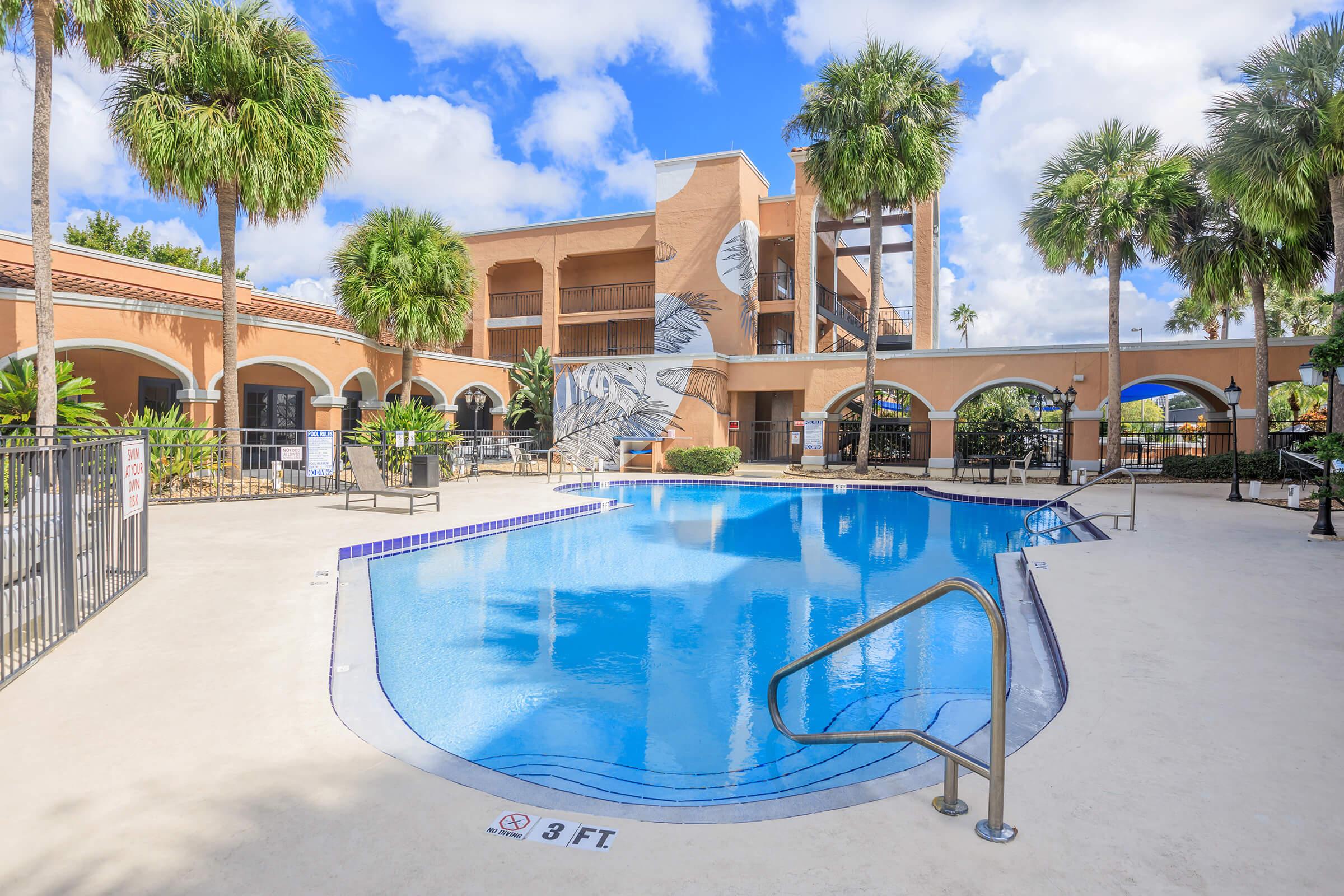 A serene outdoor pool area surrounded by palm trees and a colorful building. The pool features a shallow end marked with "3 FT." Sun loungers and a fence are visible, with partly cloudy blue skies above, creating a relaxed atmosphere perfect for leisure and relaxation.