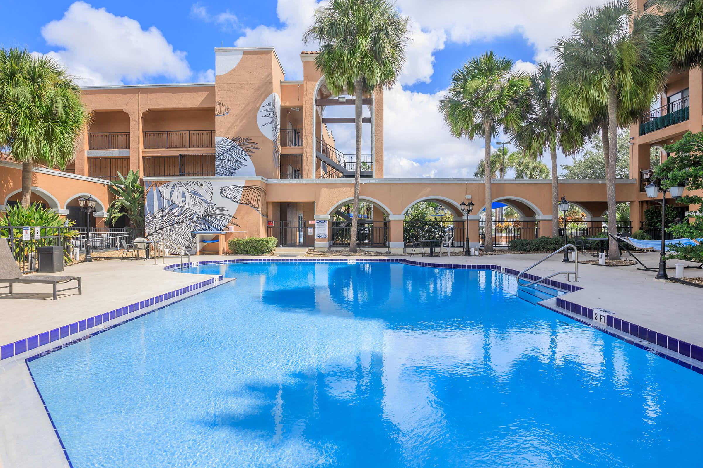 A clear blue swimming pool surrounded by palm trees and tropical landscaping, with a sunlit courtyard. The pool features a tile border and lounge chairs, and a mural of a bird can be seen on one wall of the adjacent building. Bright blue sky with fluffy white clouds overhead.