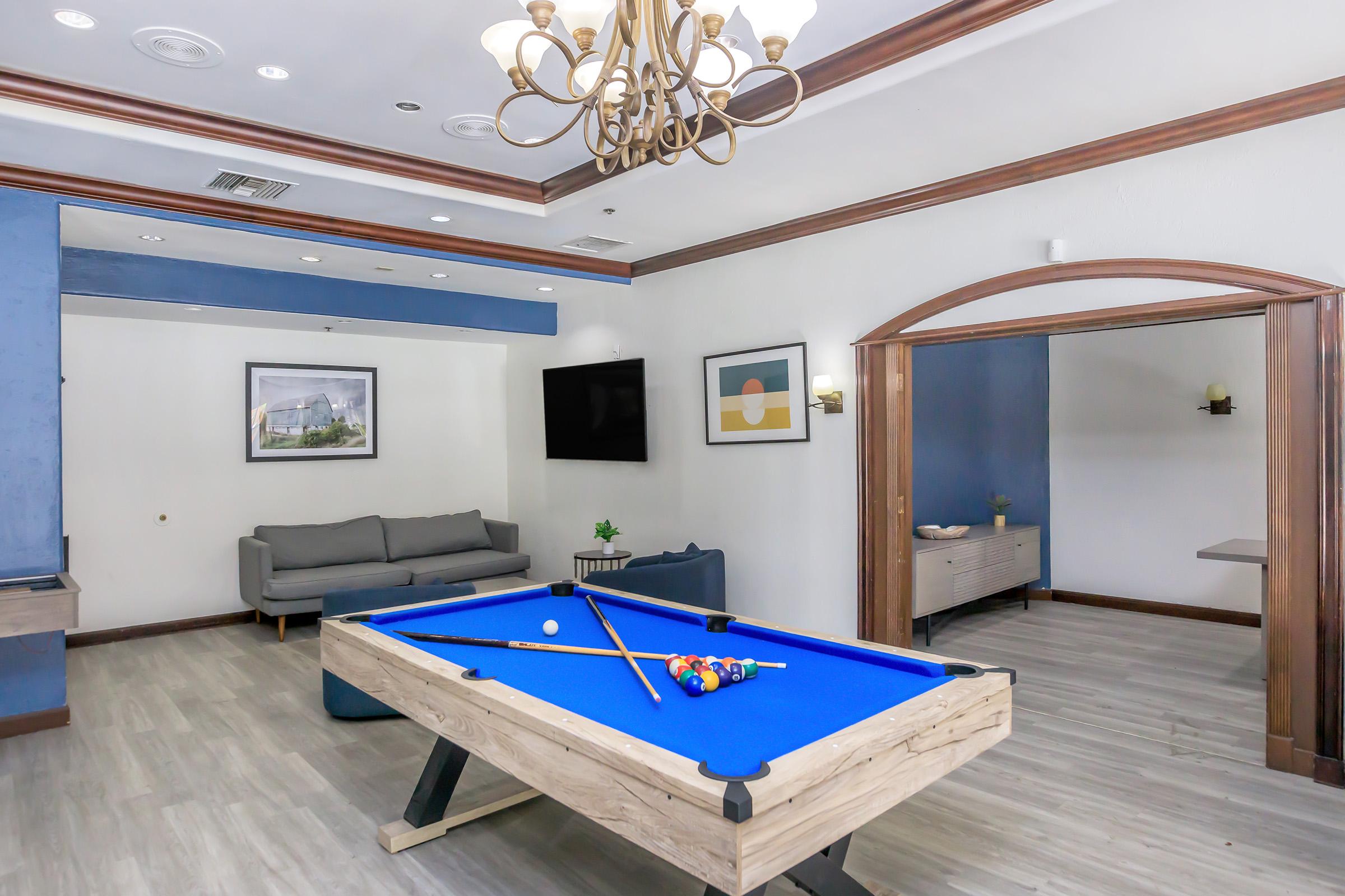 A modern recreational room featuring a blue pool table with cues and balls, a gray sofa along the wall, a flat-screen TV mounted on the wall, and framed artwork. The room has wooden accents and a stylish chandelier, providing a bright and inviting atmosphere. A separate area is visible through an archway.