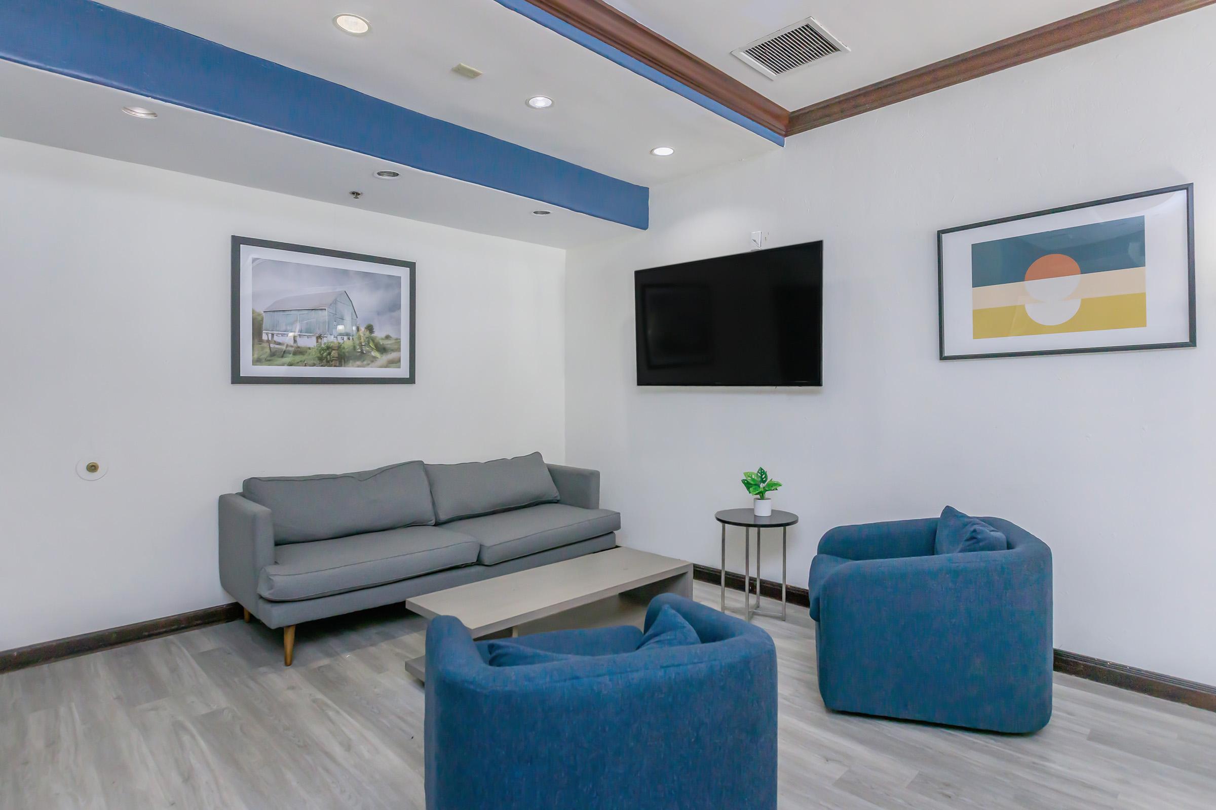 A modern, minimalist lounge area featuring a gray sofa, two blue armchairs, and a small round table. On the walls, there are two framed artworks, and a flat-screen TV is mounted above the sofa. The space has neutral-colored walls and a light wooden floor, creating a welcoming atmosphere.