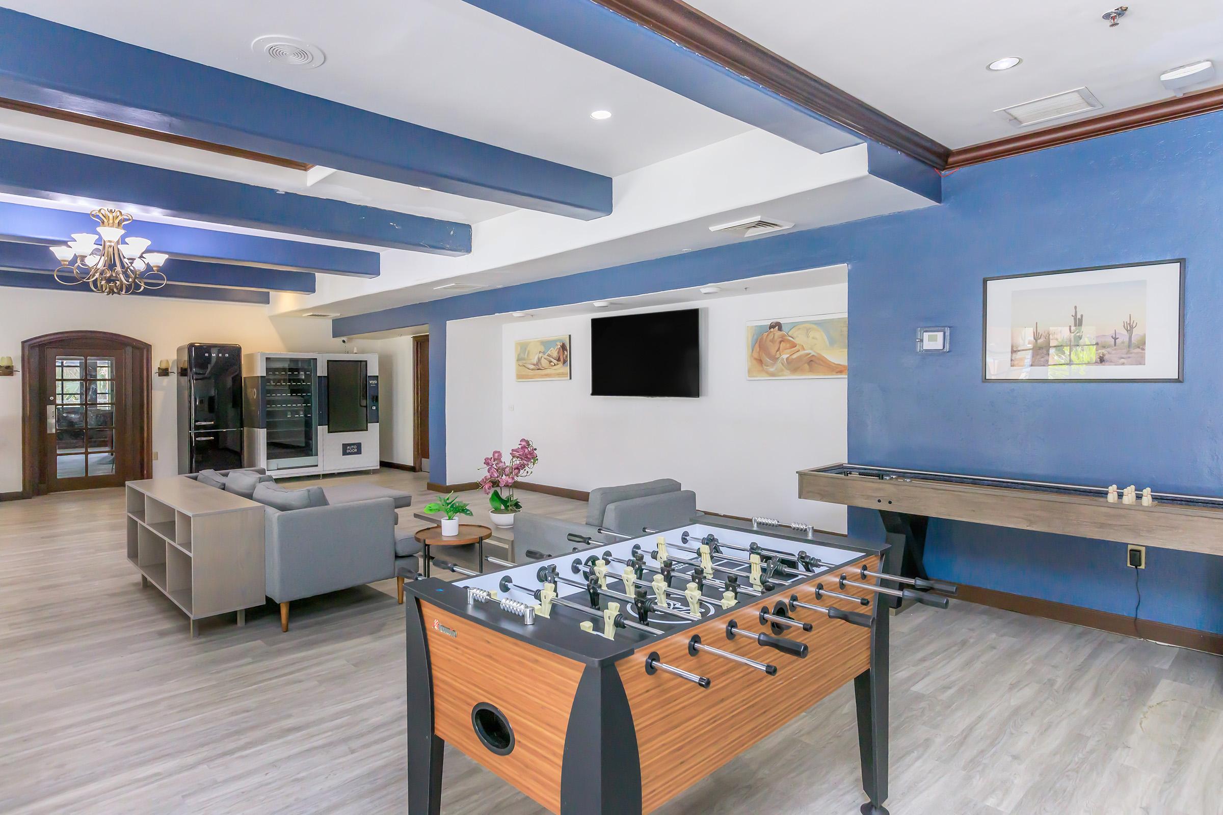 A modern recreation room featuring a foosball table, a comfortable seating area with a gray sofa, a wall-mounted TV, and a ping pong table. The room is decorated with blue walls, wooden beams, and a chandelier, creating a vibrant and inviting atmosphere.