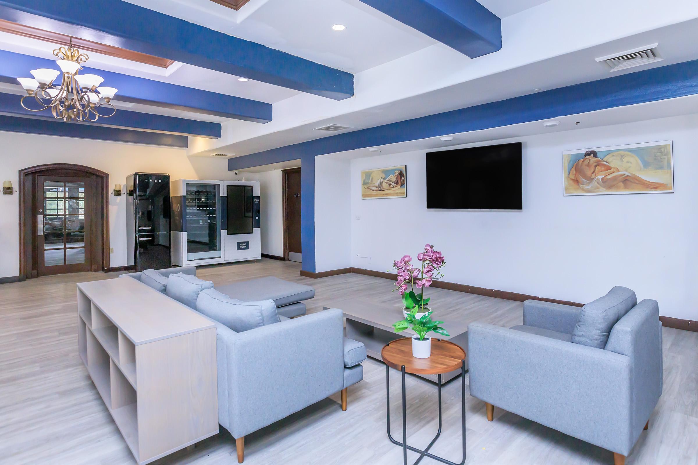 A modern lounge area featuring gray sofas and a wooden coffee table. There is a large flat-screen TV on the wall, along with two decorative paintings. The space has blue beams on the ceiling, soft lighting from a chandelier, and a small plant centerpiece on the table. A vending machine is visible in the background.